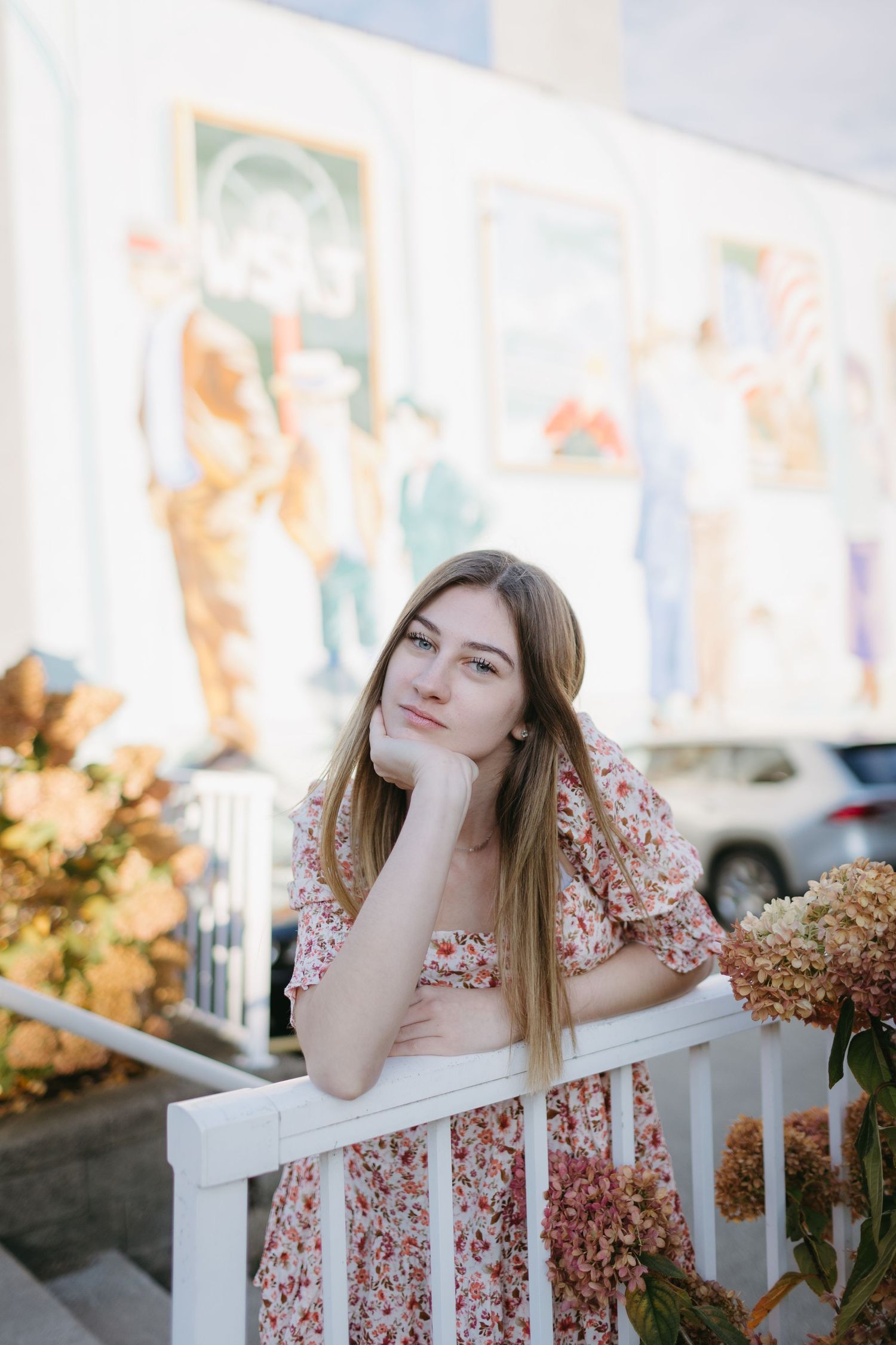 Young woman in floral dress leaning on white railing with autumn foliage in background.