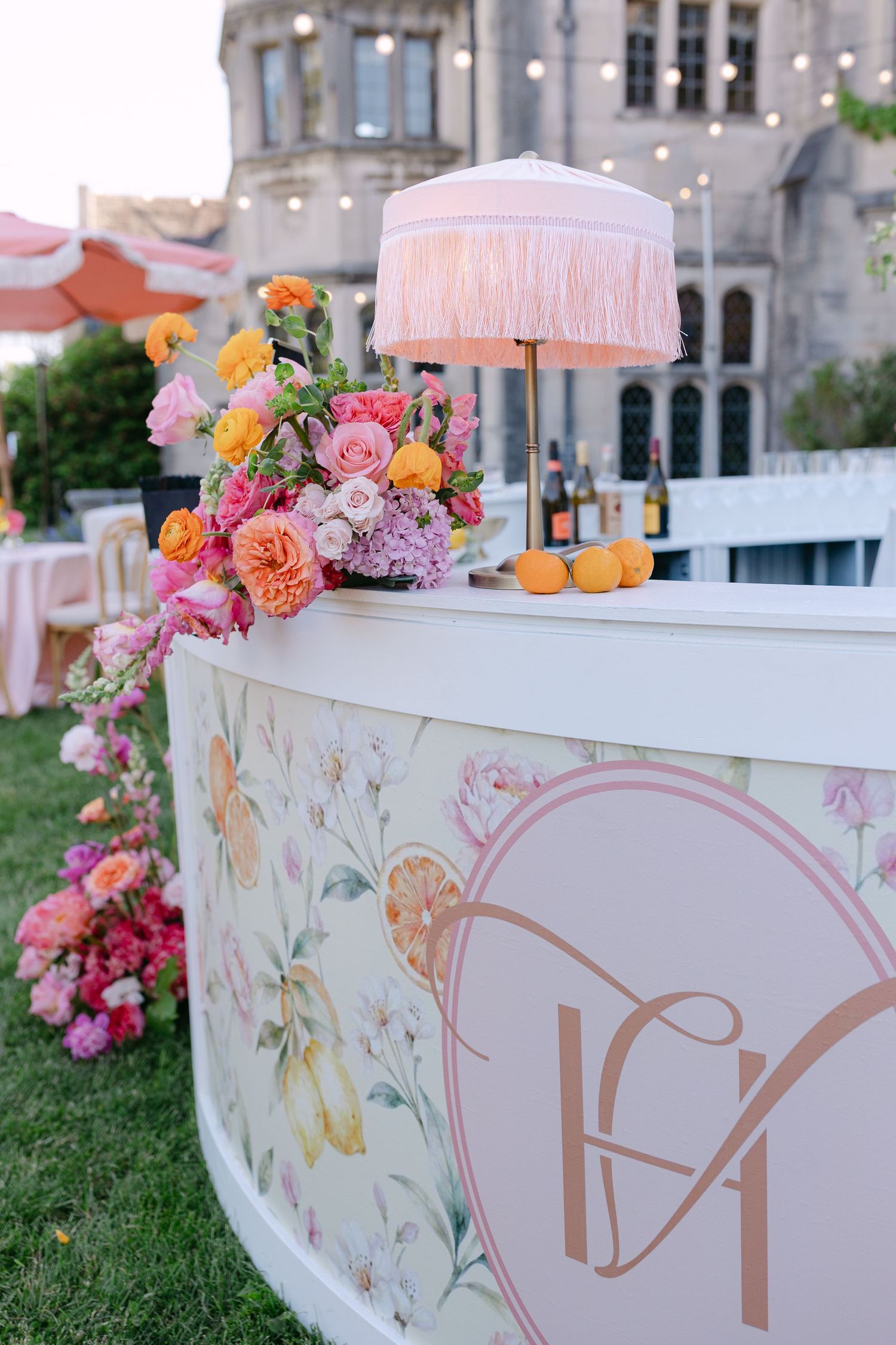 Elegant outdoor bar with floral decor, pink fringed lamp, monogram logo, and colorful roses at a garden wedding event.