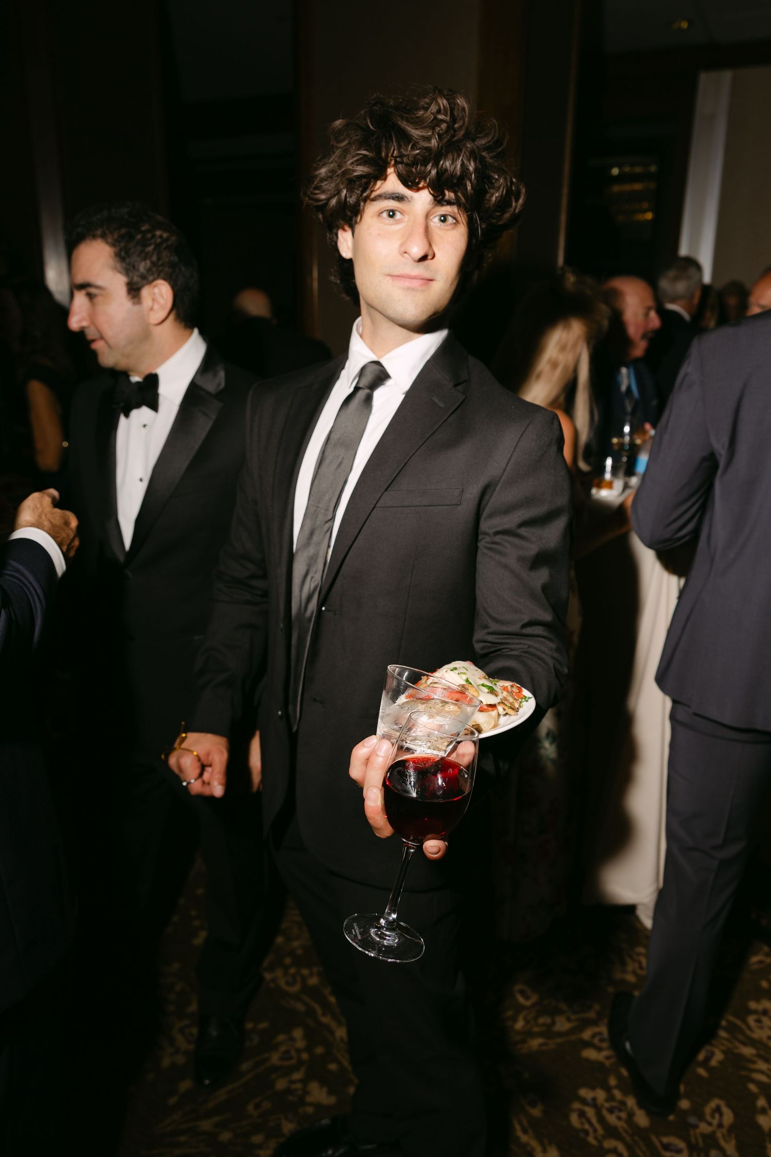 A young man in a black suit and tie holds a cocktail glass at a formal evening event.