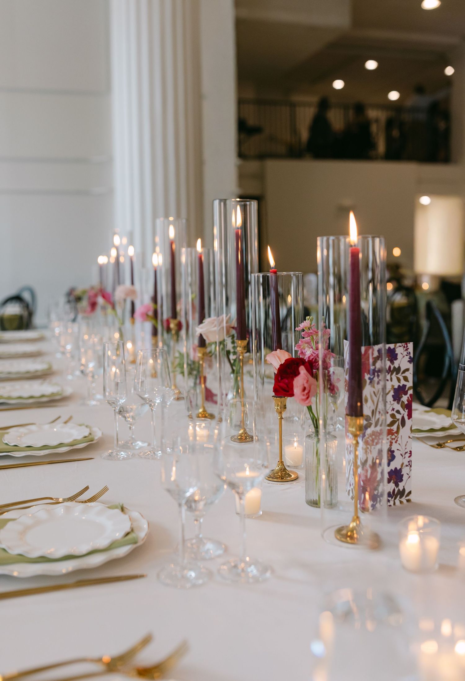 Elegant wedding table setting with tall glass candle holders, burgundy taper candles, gold accents, and floral arrangements.