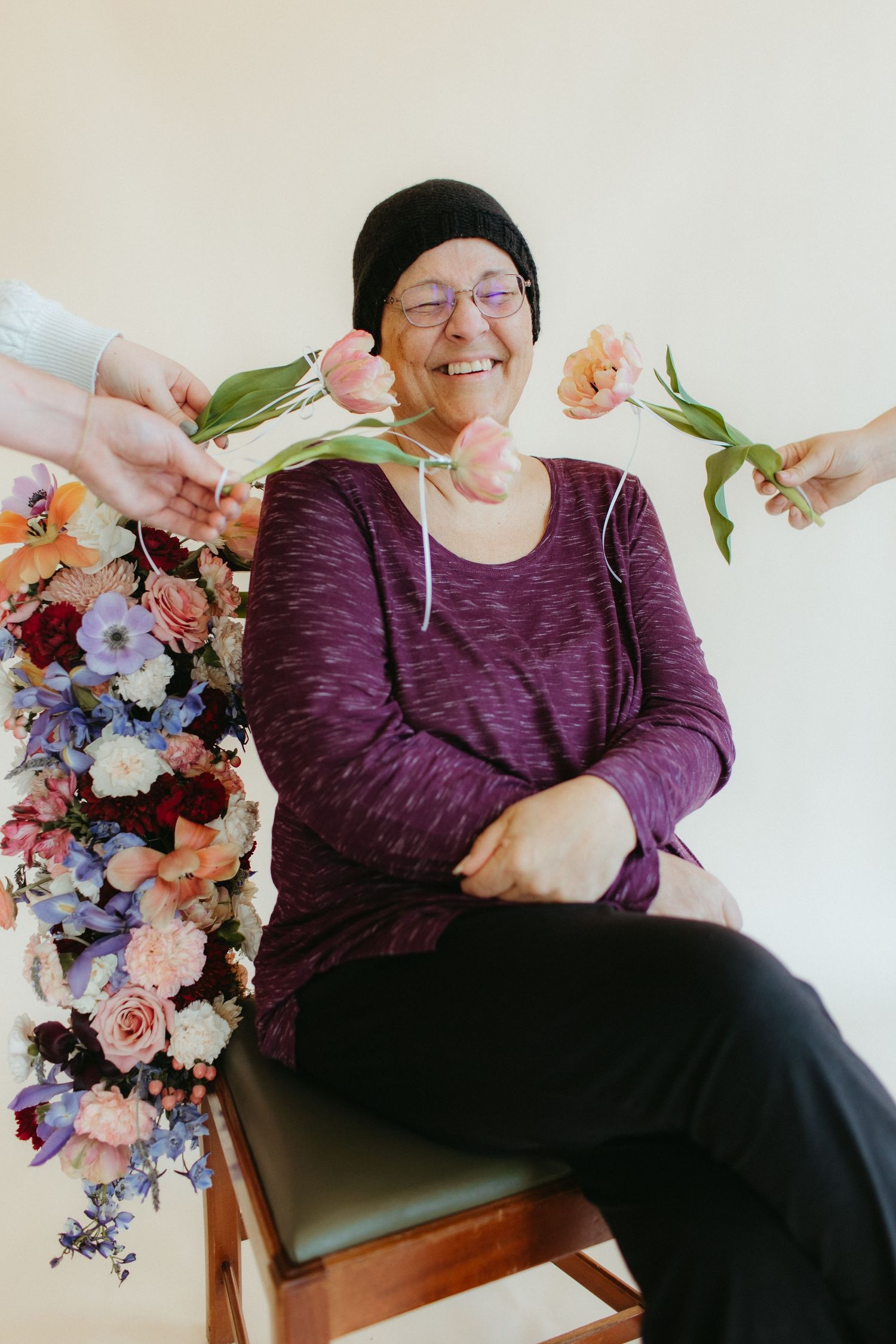 A person in purple wearing a beanie smiles while hands offer flowers from a colorful floral arrangement.
