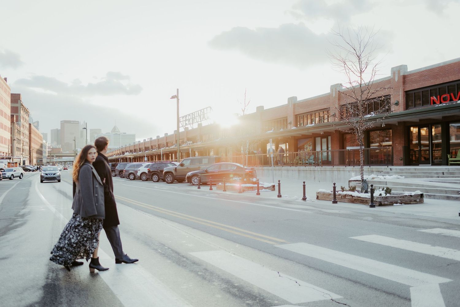 Couple standing on snowy sidewalk at sunset in front of urban storefronts.