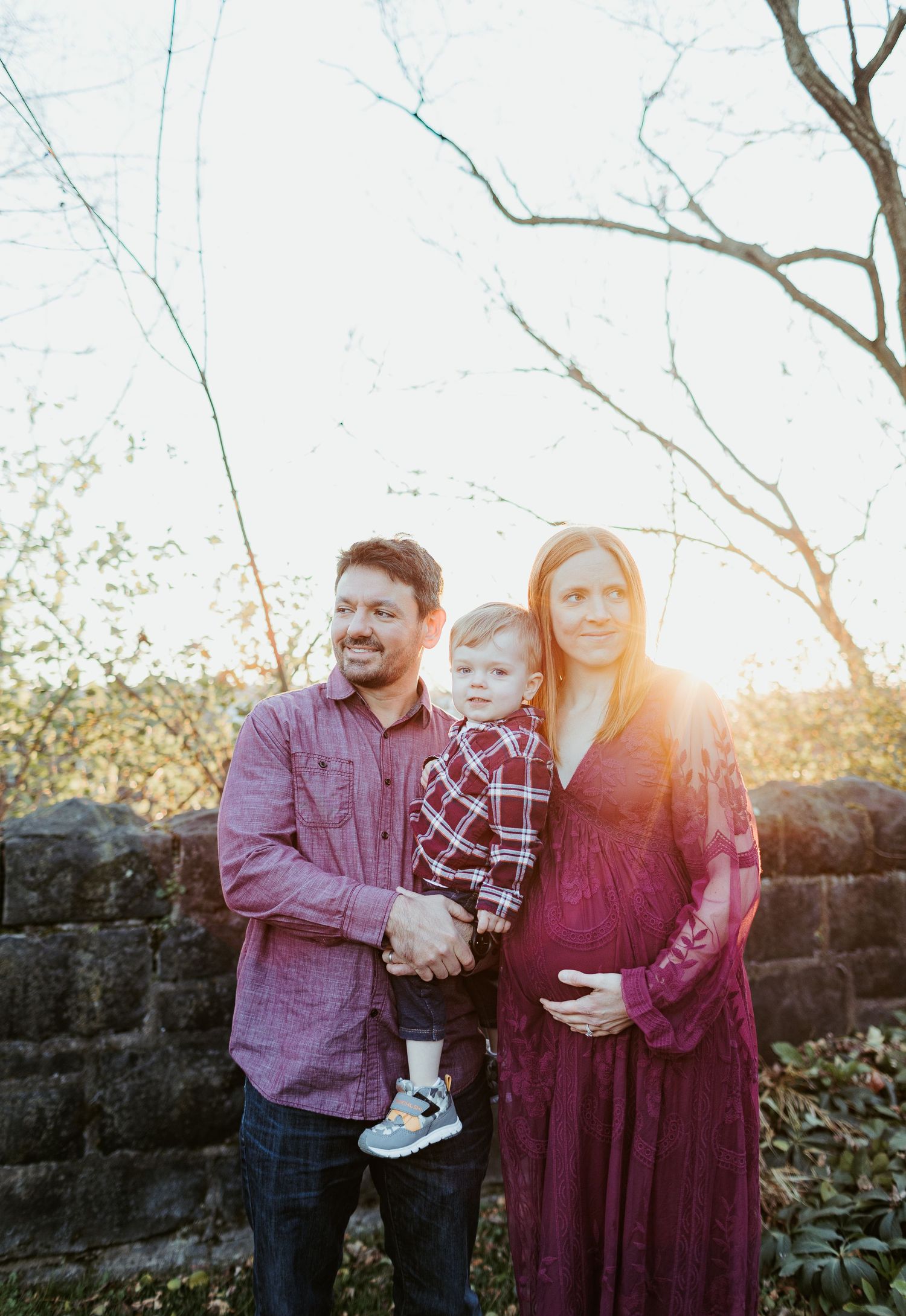 A family stands together at sunset against a stone wall backdrop with bare tree branches overhead.