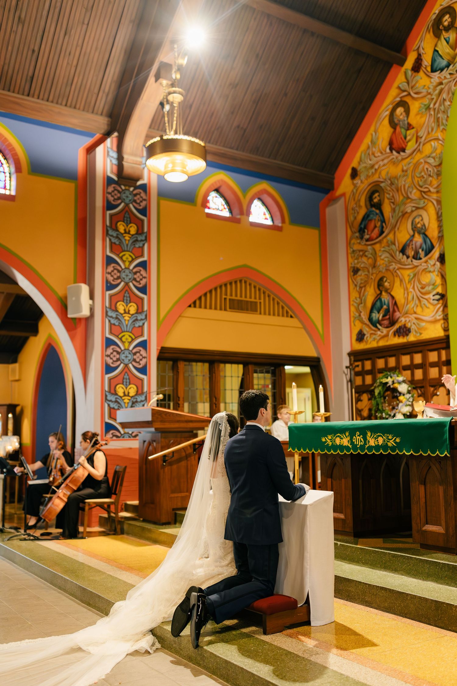 A wedding couple kneels at the altar in a colorful church with ornate decorative walls and arched architecture.