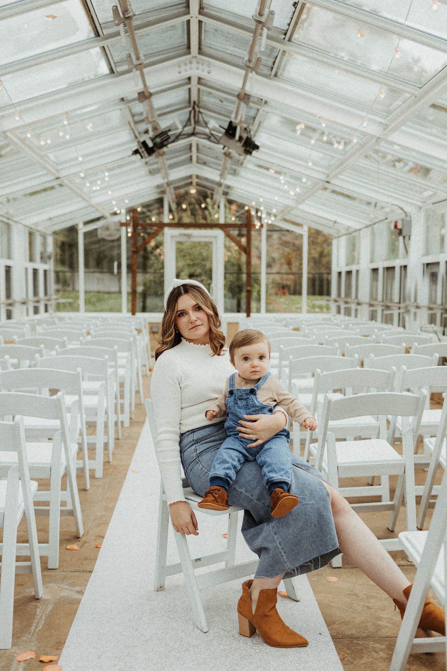 A parent and child sit together on white chairs in an elegant glass greenhouse wedding venue.