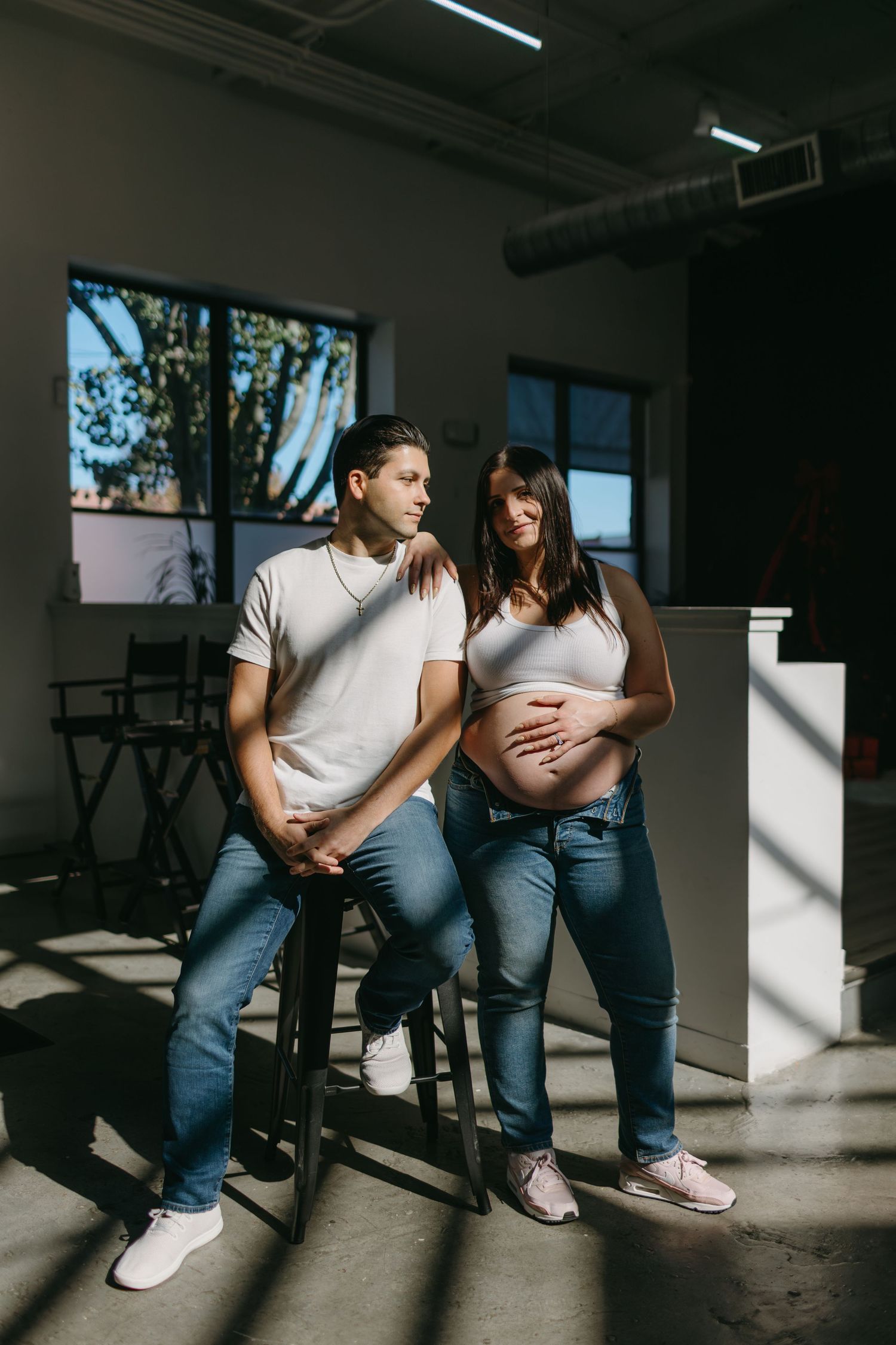 Expecting couple in casual denim and white shirts posing in sunlit industrial space.