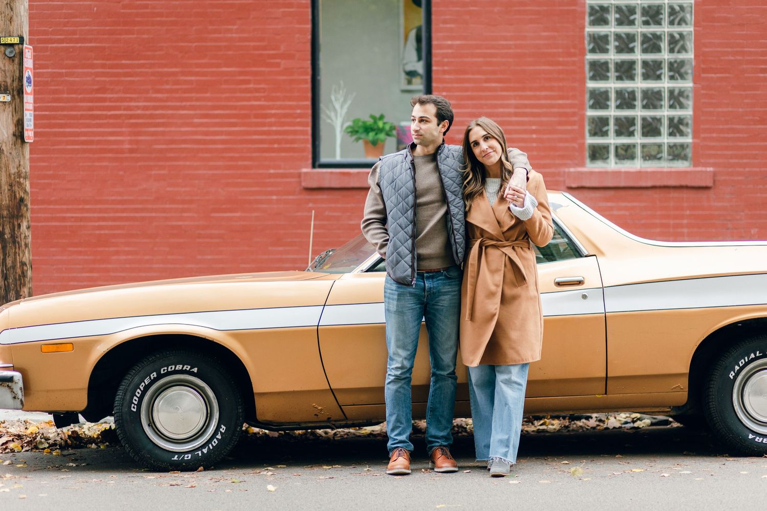 Couple leaning against vintage gold car in front of red brick building.