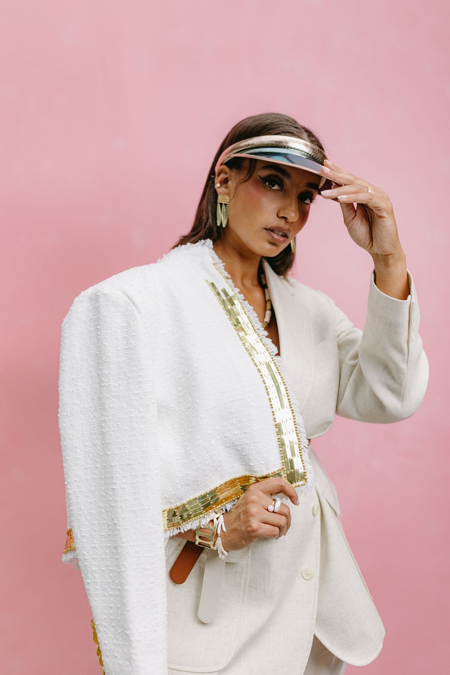 Fashion model in a white blazer with gold trim and clear visor poses against pink background.
