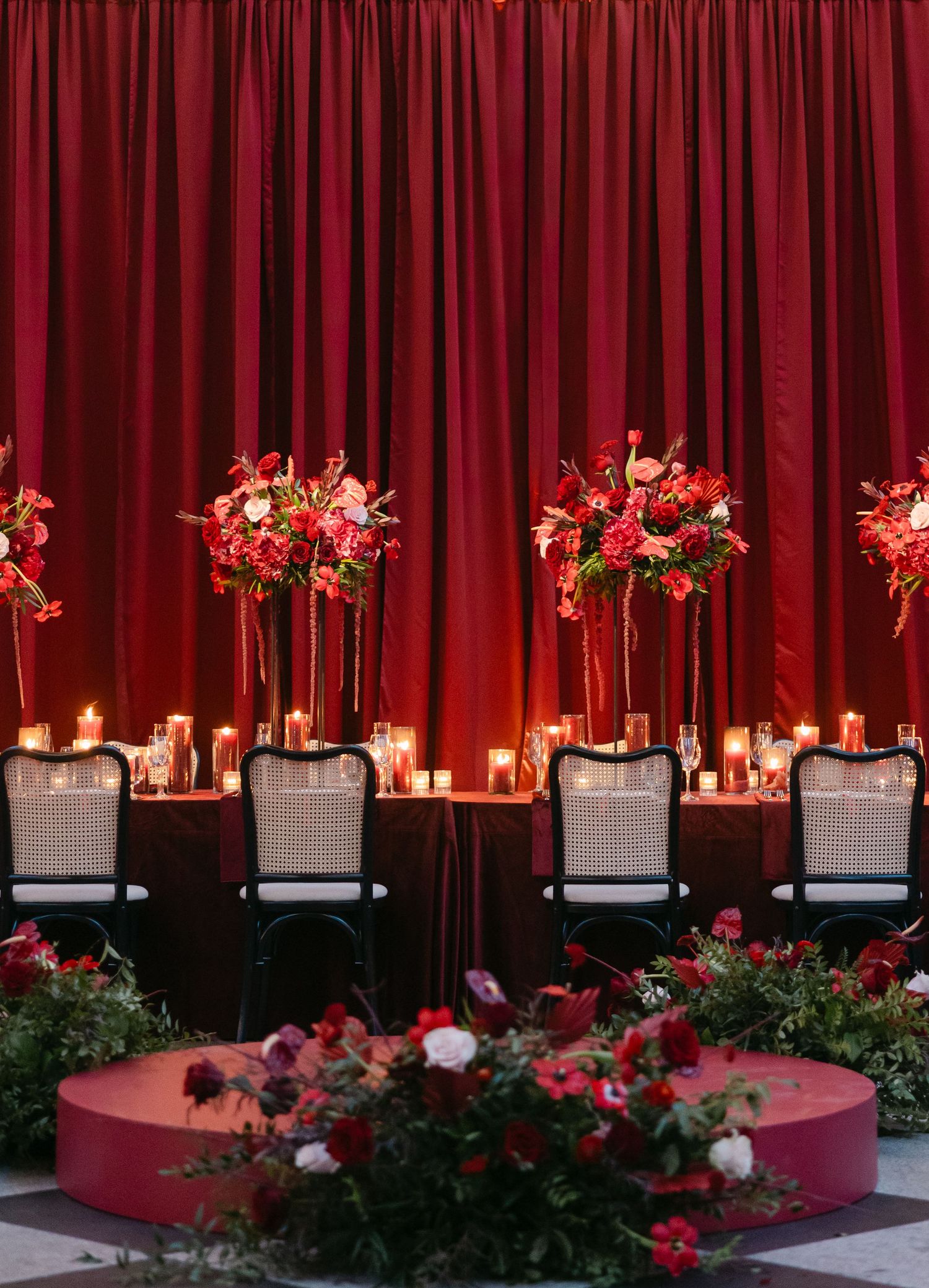 Elegant wedding reception table with tall red floral centerpieces against a dramatic red velvet curtain backdrop and candlelight.