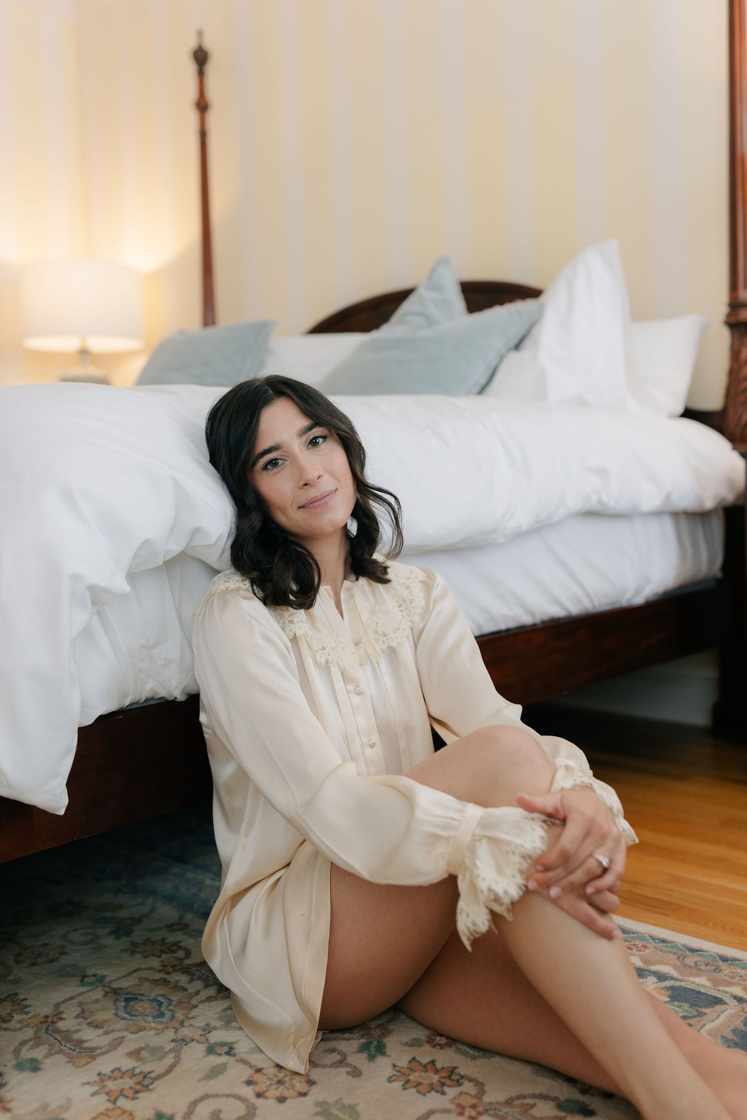A person sits on a hardwood floor beside a bed wearing a cream-colored flowy dress shirt in a sunlit bedroom.