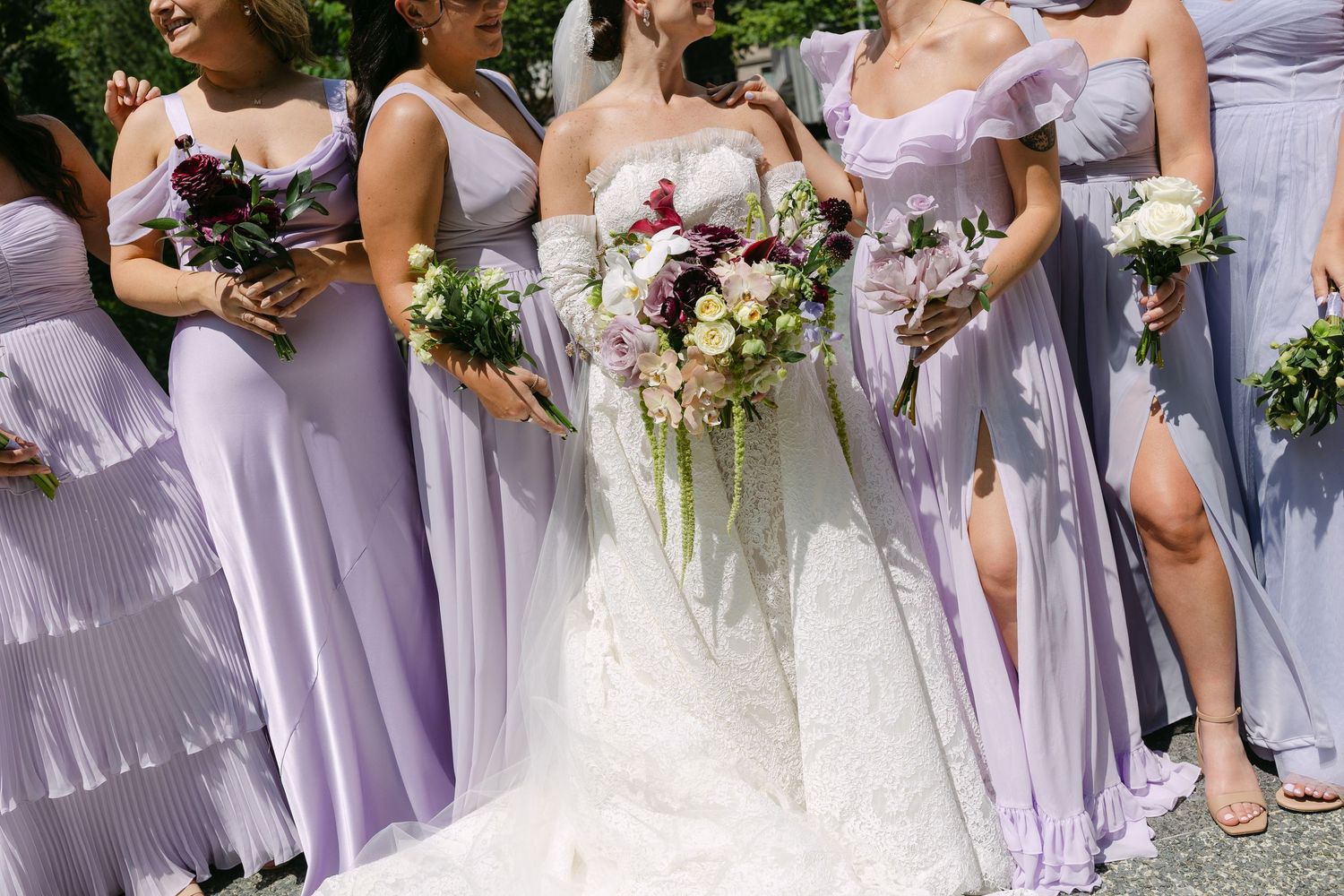 A wedding party stands together in lavender bridesmaid dresses holding floral bouquets alongside a bride in white.