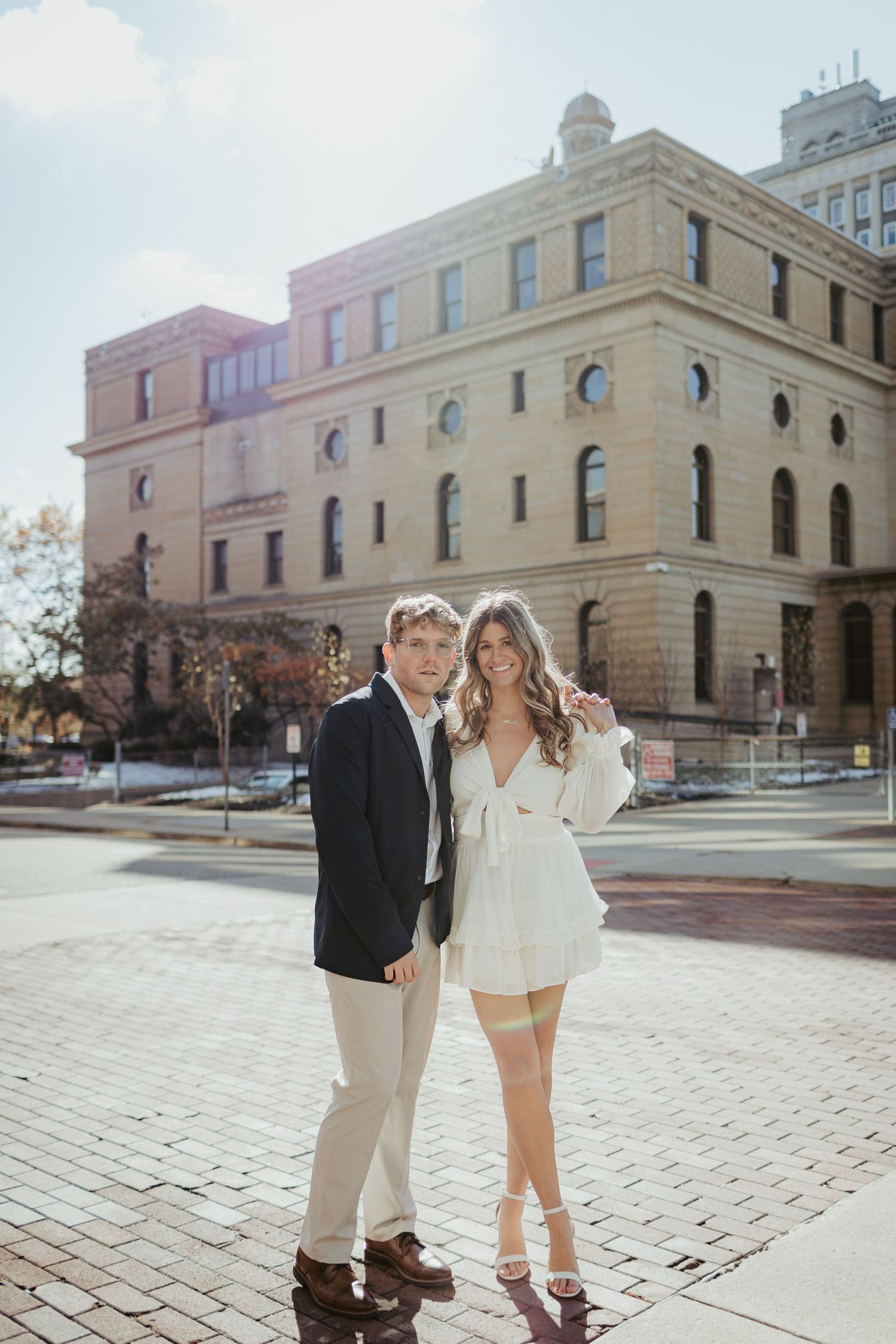 Couple dressed in formal attire posing in front of a historic building on a sunny day.