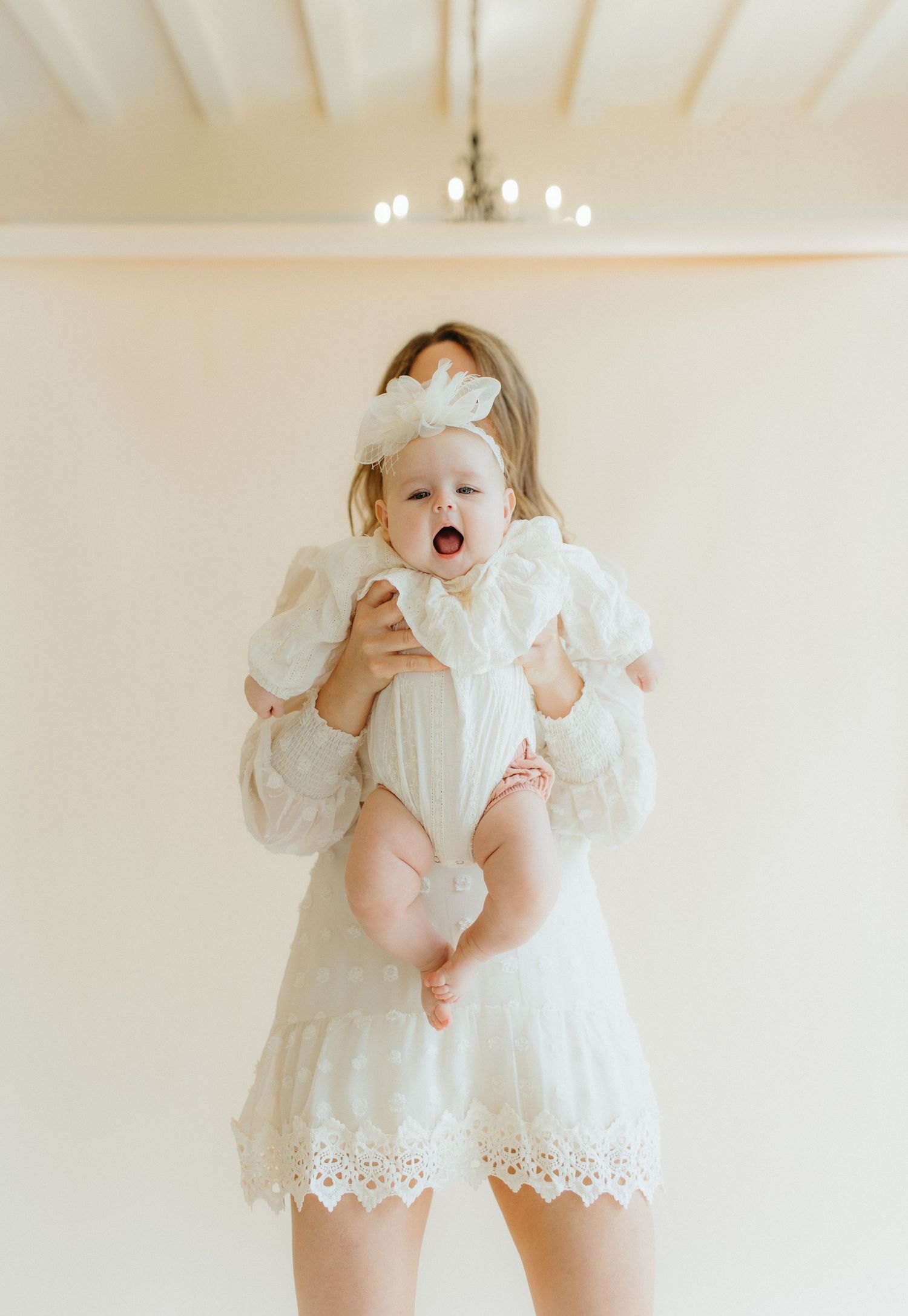 Baby wearing white dress held up high against cream colored wall with chandelier.