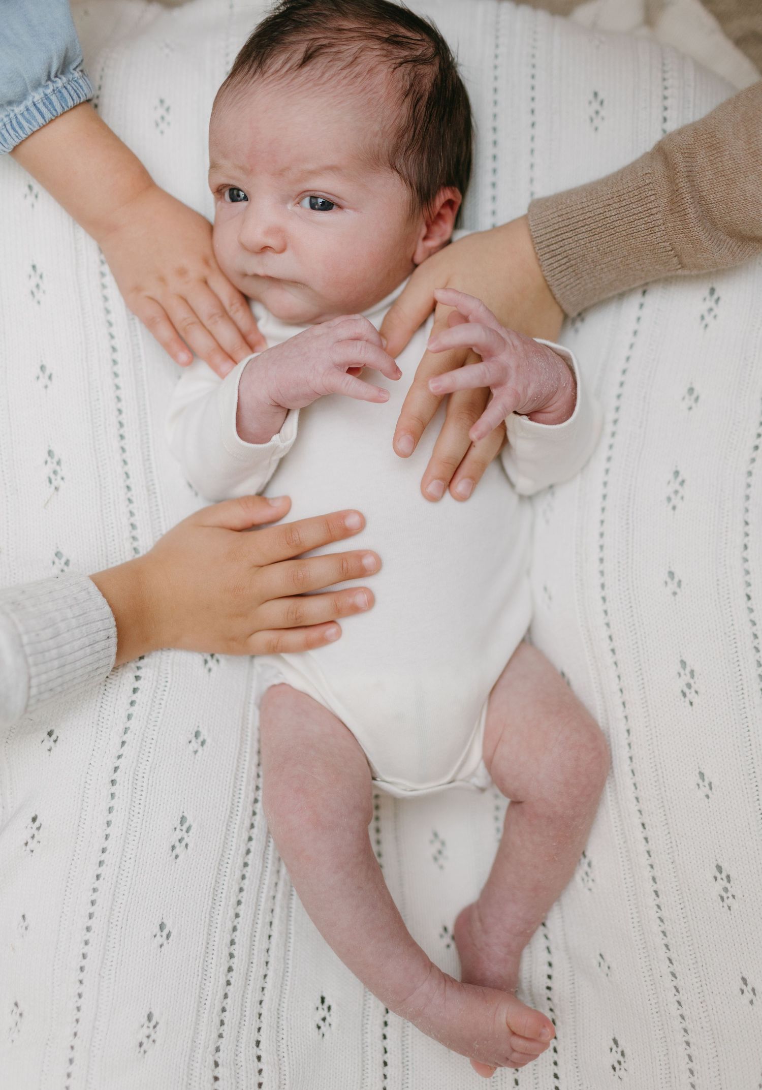 A baby in a white onesie lying on a textured white blanket with hands reaching to touch.