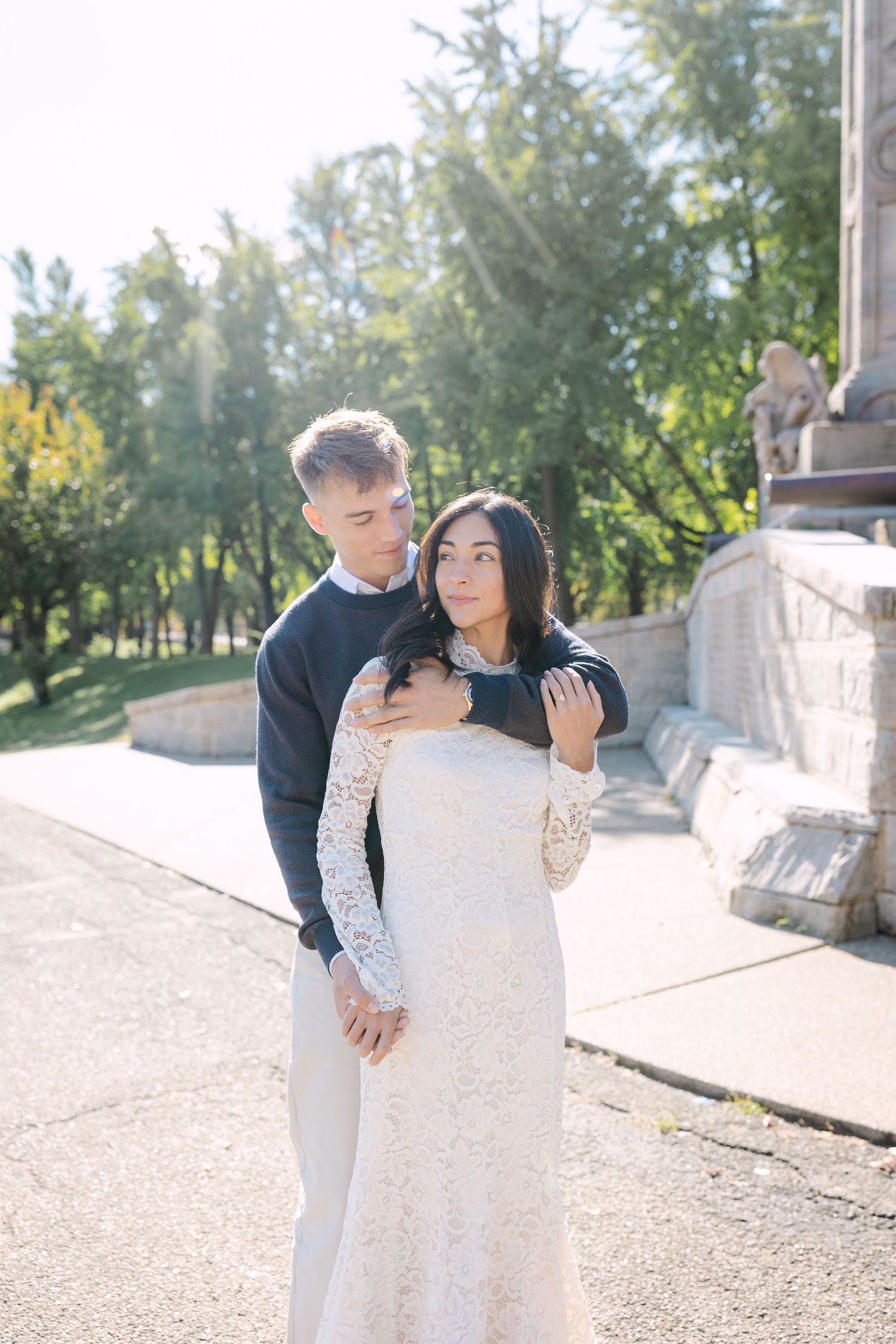 A couple in formal attire embraces lovingly on a sunlit path surrounded by trees and greenery.