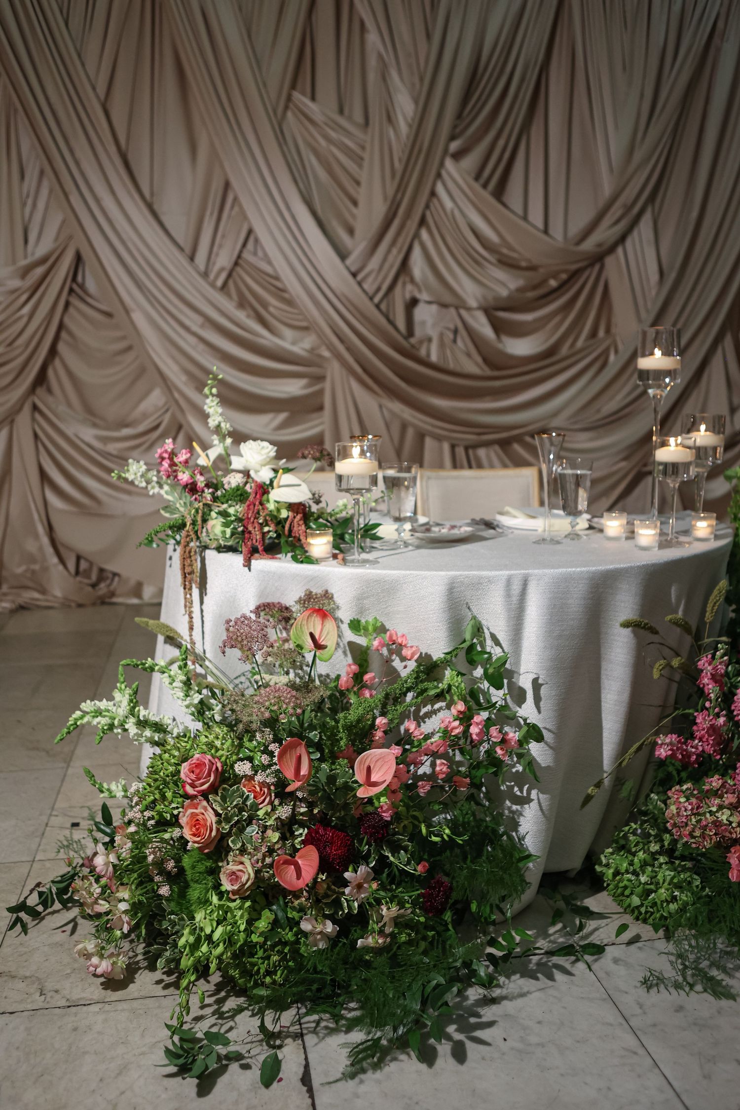 Elegant wedding sweetheart table with floral arrangements, candles, and draped fabric backdrop.