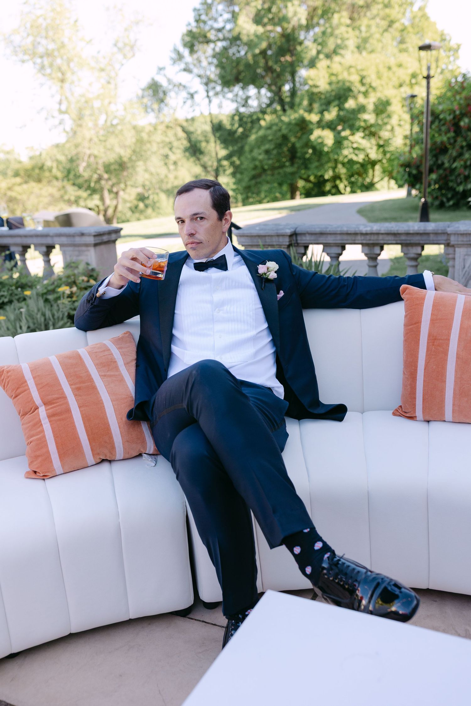 Groom in navy tuxedo holding whiskey glass on white outdoor sofa with orange pillows at wedding reception.