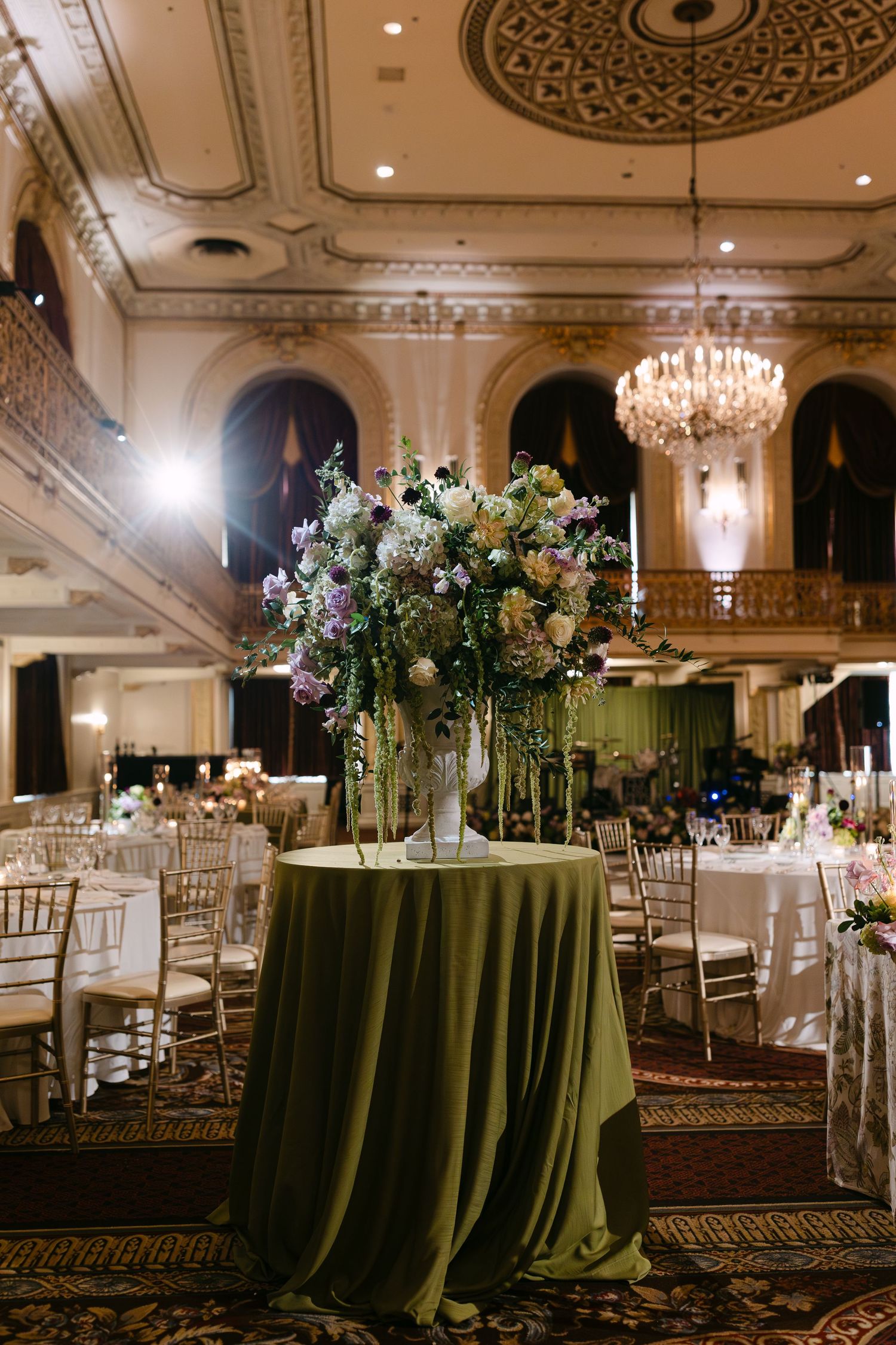 Elegant wedding reception table with tall floral centerpiece and sage green tablecloth in ornate ballroom with crystal chandelier.