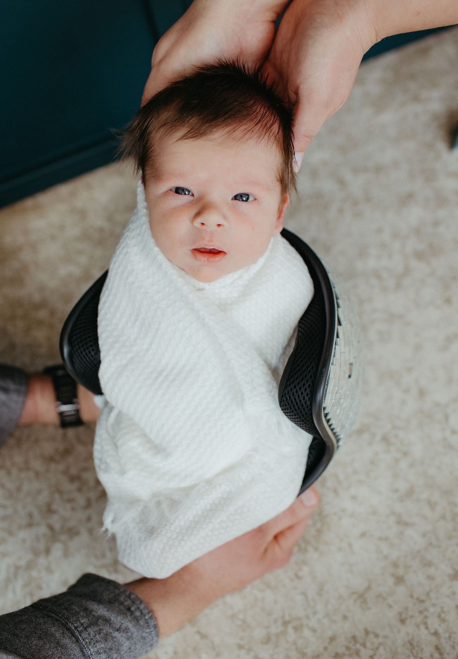 Baby in white sweater held by hands against carpeted background.