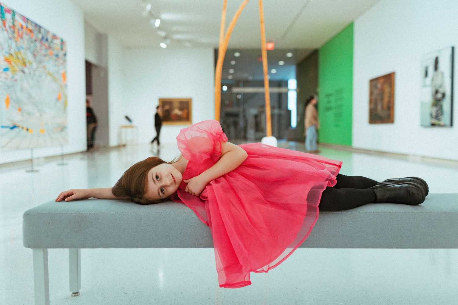 Child in pink dress lying playfully on gray bench in modern art gallery.