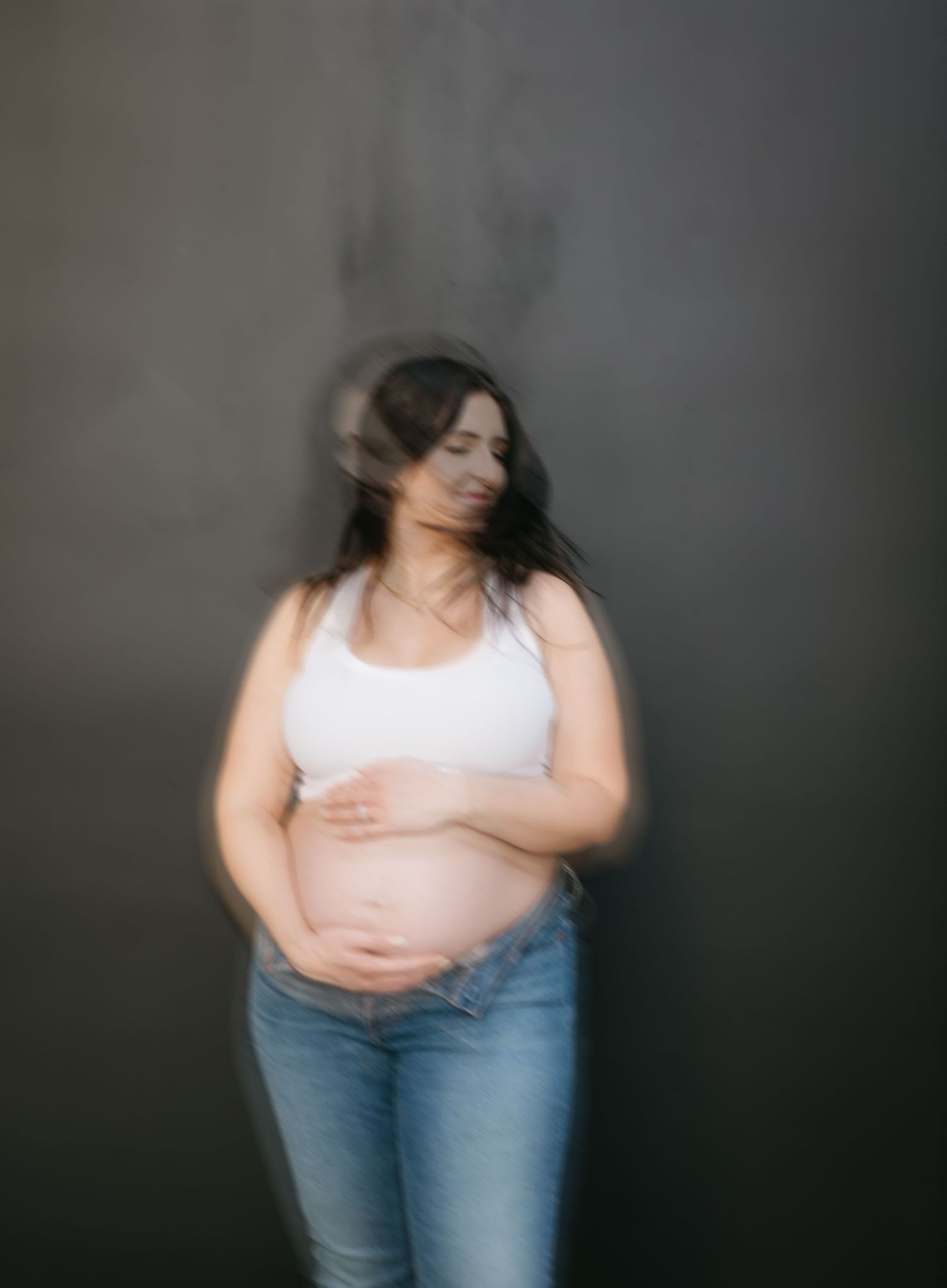 Artistic blurred maternity portrait of a pregnant woman in white top and jeans against dark background.