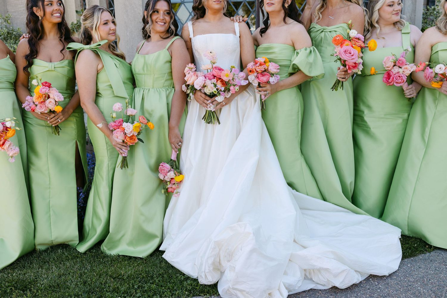 Bride in white gown with bridesmaids in sage green dresses holding colorful pink and orange bouquets outdoors.