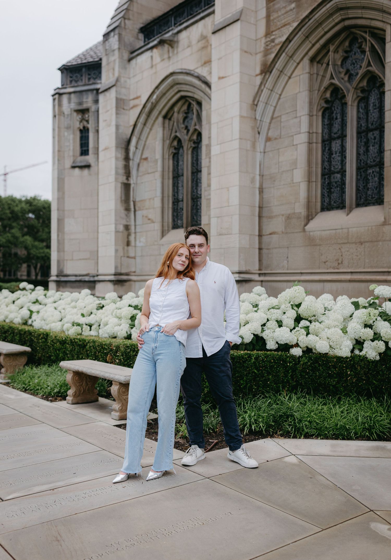 A couple sits together on stone church steps surrounded by white hydrangea bushes on a summer afternoon.
