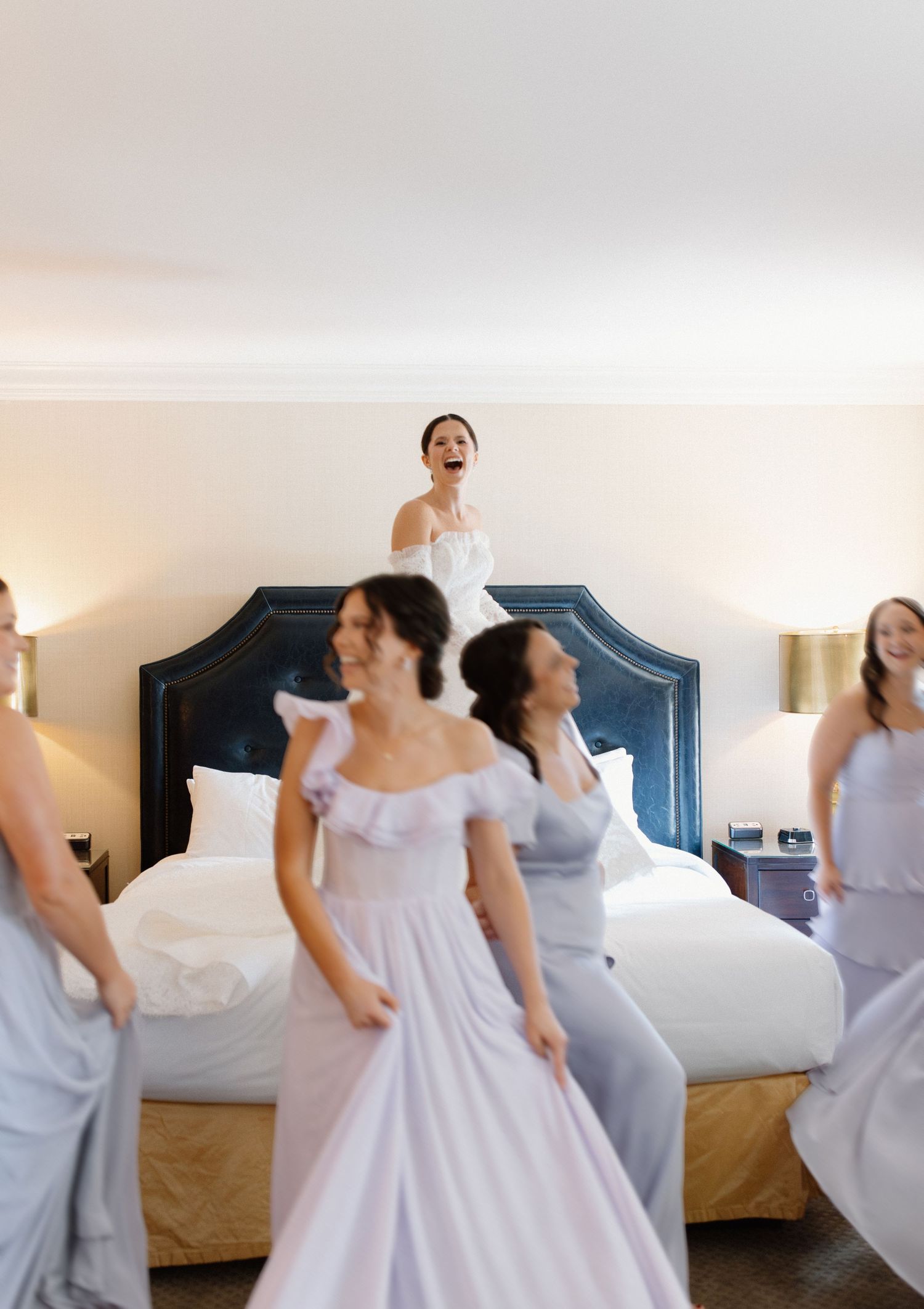 Bridesmaids in flowing white dresses laugh and dance together in a hotel room with a navy blue upholstered headboard.