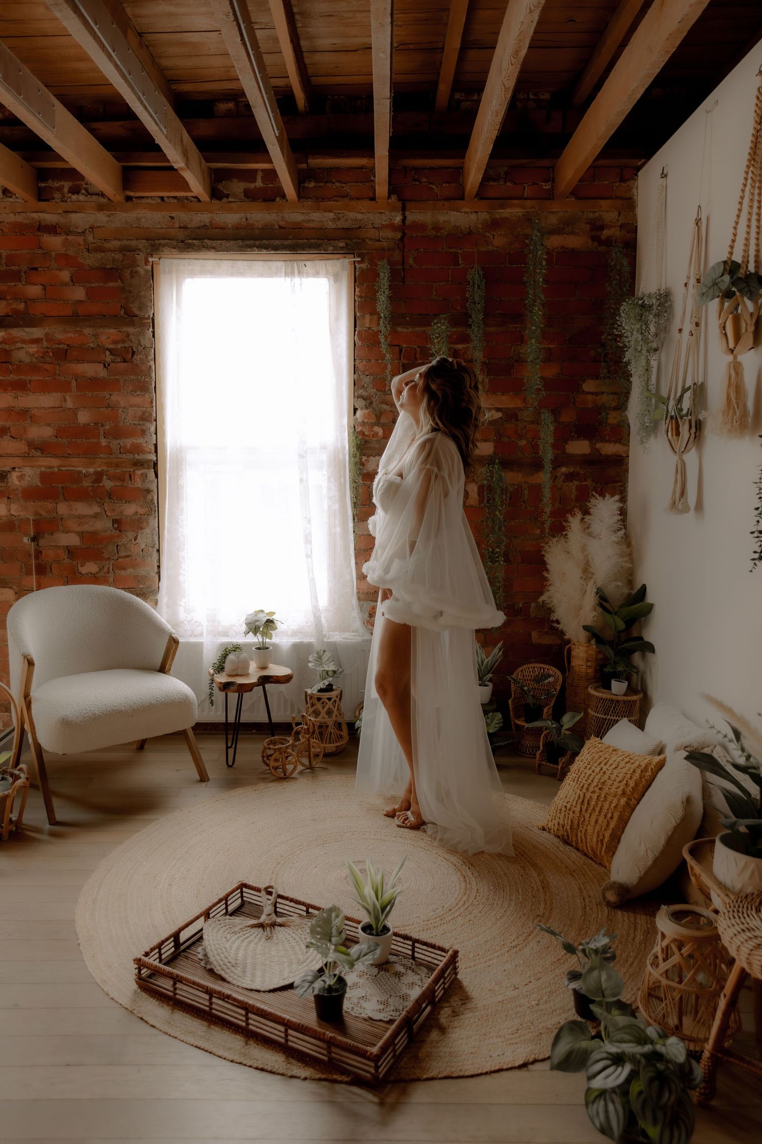 A flowing white dress billows near a bright window in a rustic brick room with hanging plants and boho decor.