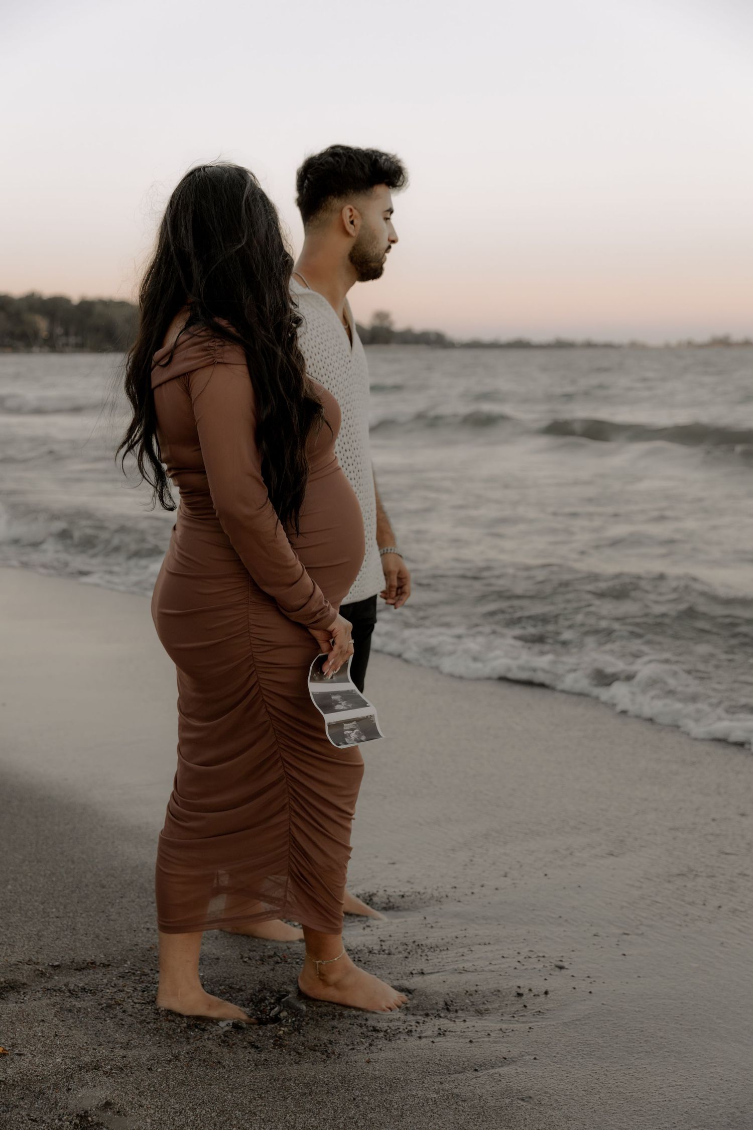 An expectant couple standing on a beach at sunset holding an ultrasound photo.