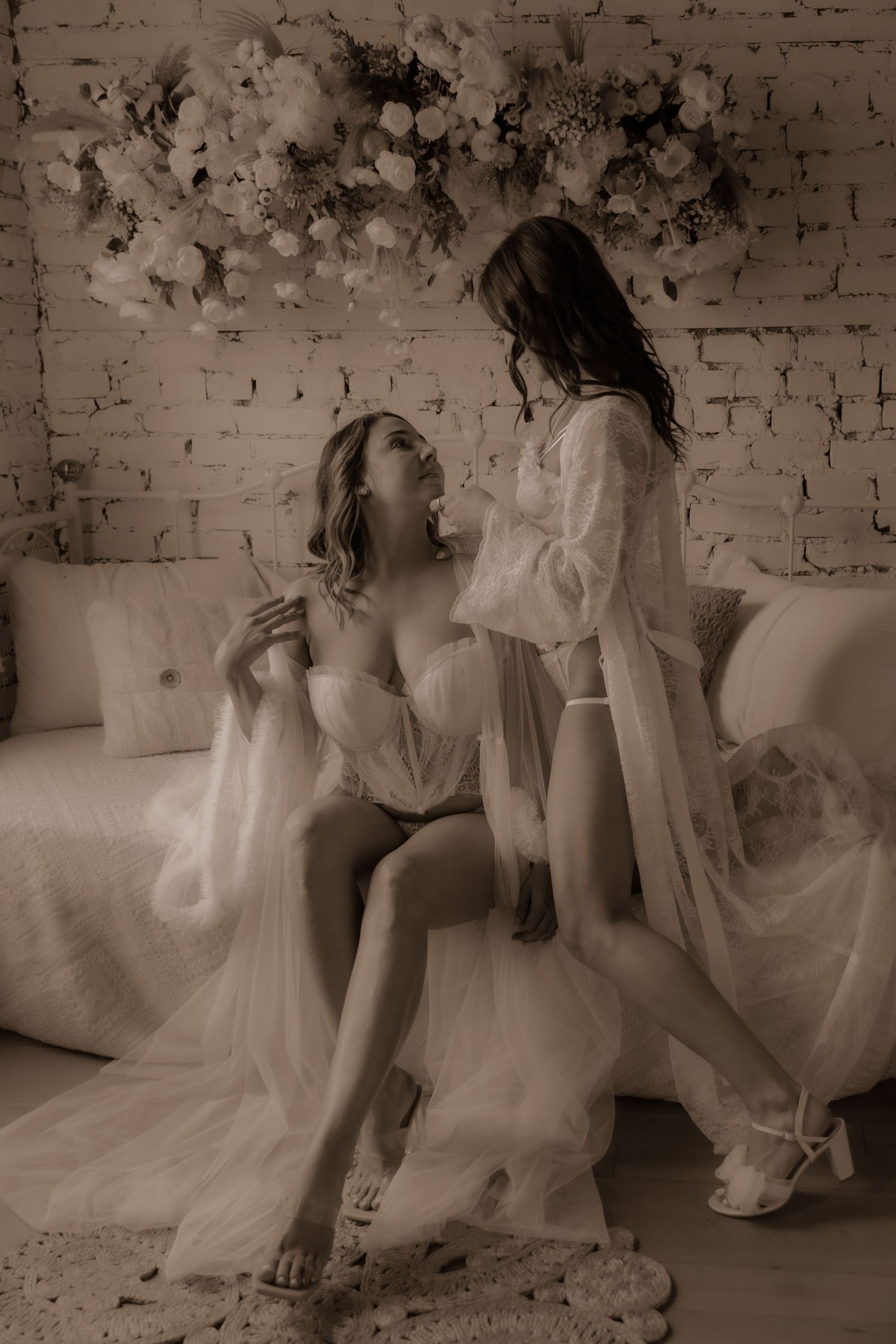 Black and white boudoir photo with two models in white lace against exposed brick wall.