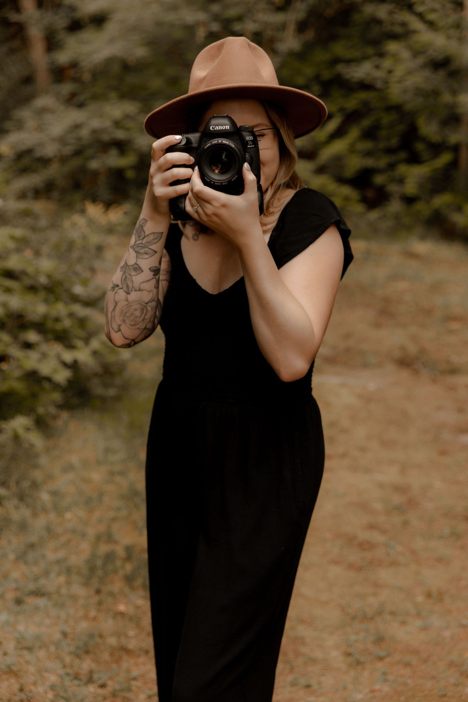 Person in black maxi dress and brown hat photographs with professional camera outdoors.