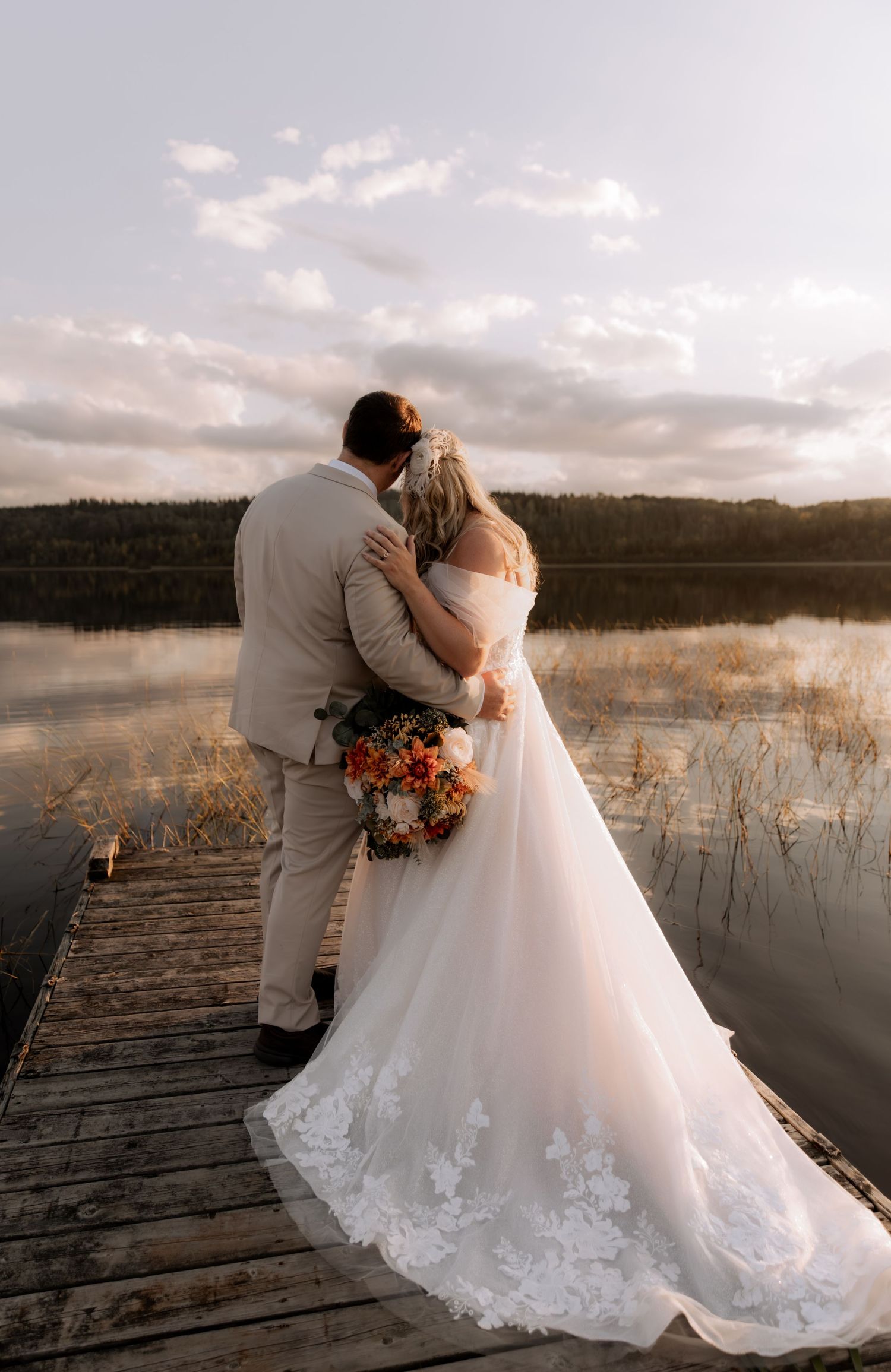 A couple embraces on a wooden dock at sunset, with the bride wearing a flowing lace dress and holding a vibrant bouquet.