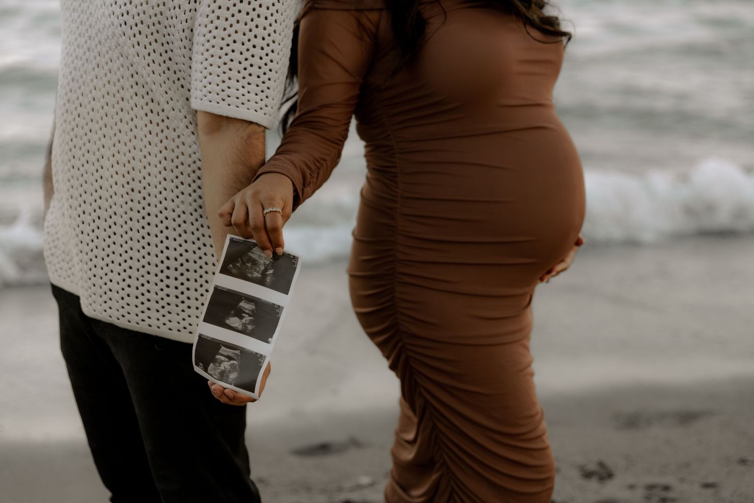 Close-up of pregnant woman in brown dress holding ultrasound photos beside partner.