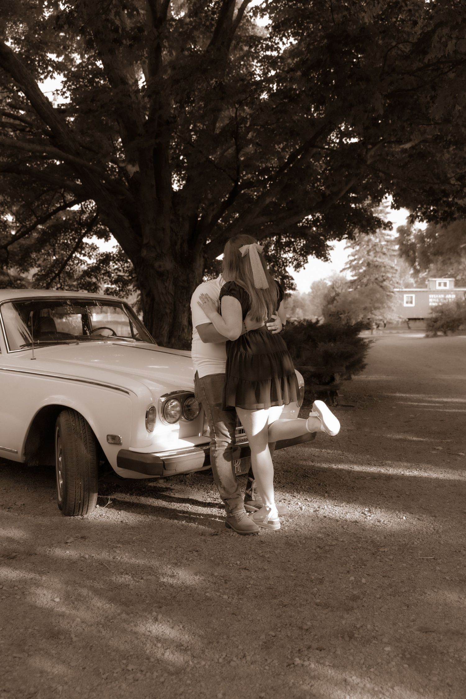A romantic vintage-style photo of a couple by a classic car under a large oak tree.