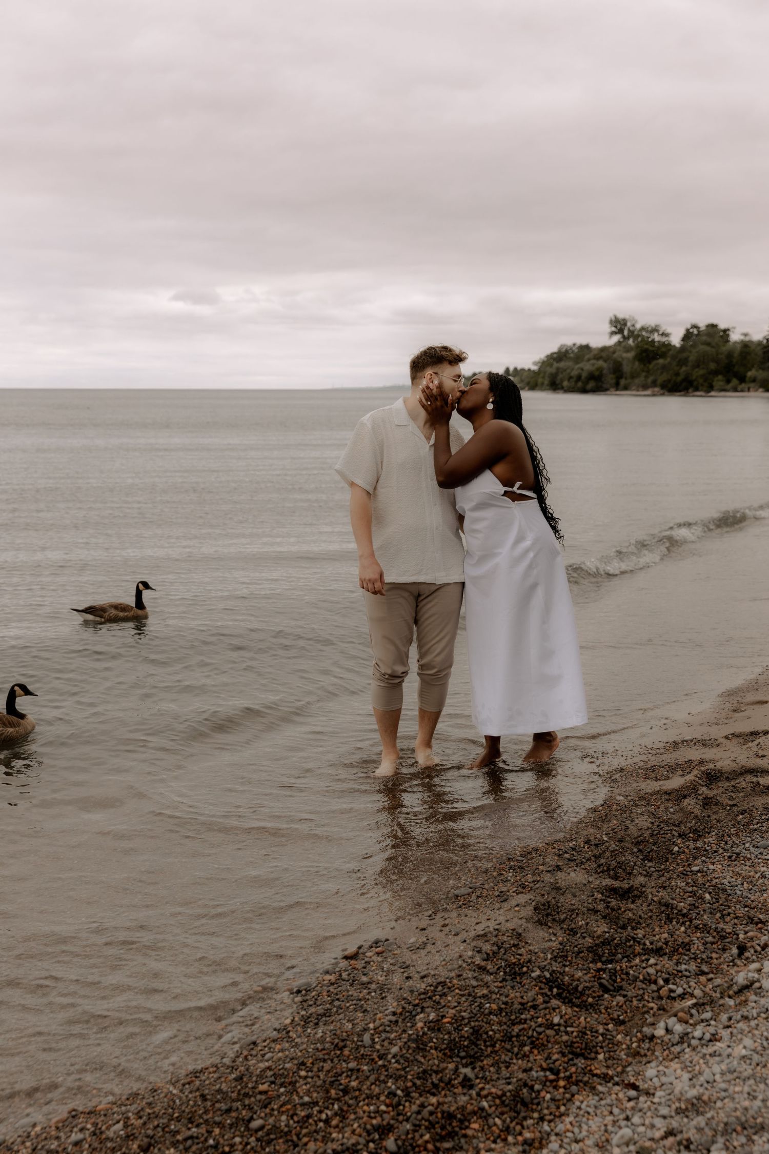 A couple shares an intimate moment on the beach with ducks swimming in the background.