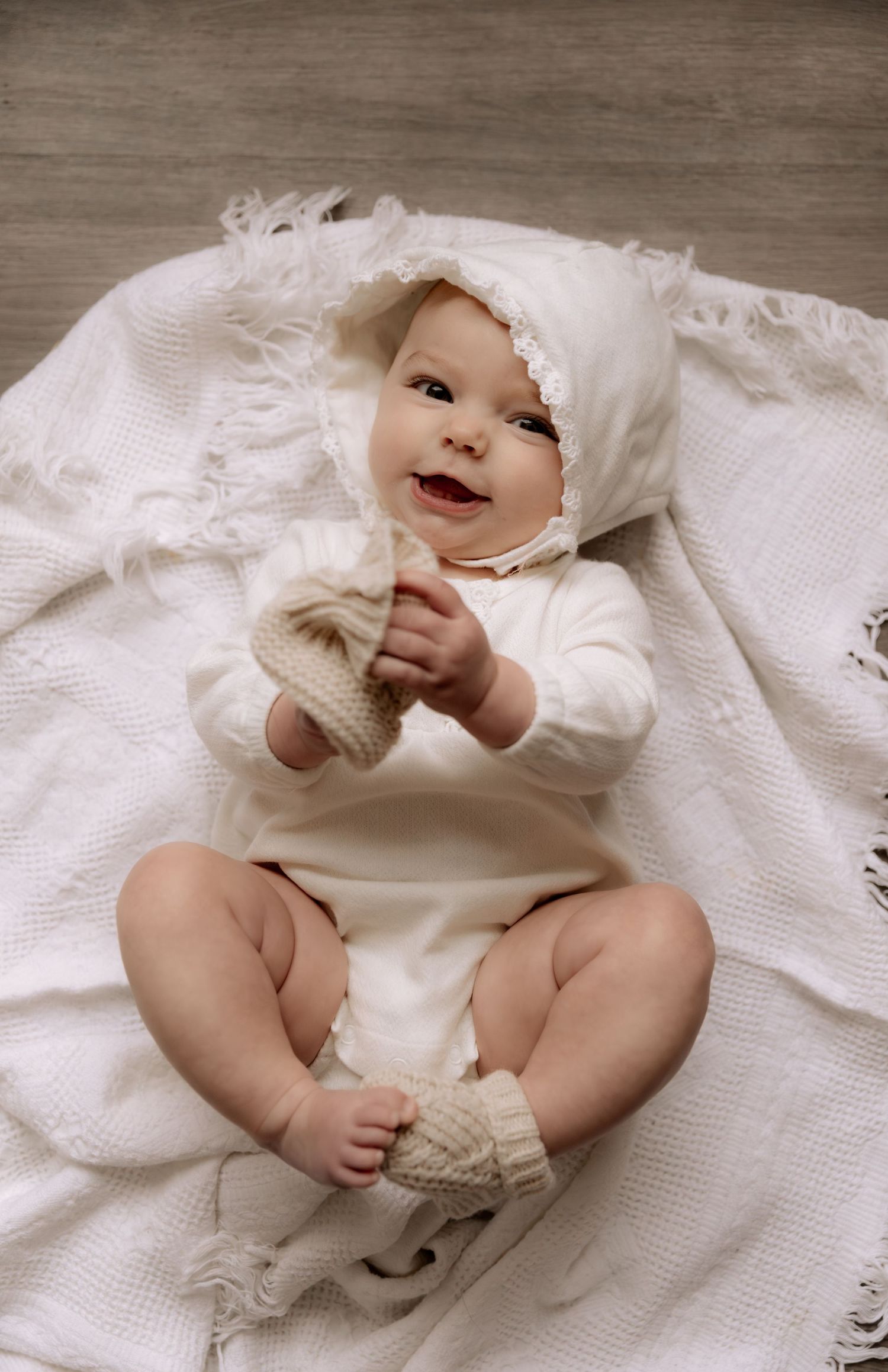Smiling baby in white onesie, lace bonnet, and knit booties lying on a soft white blanket.