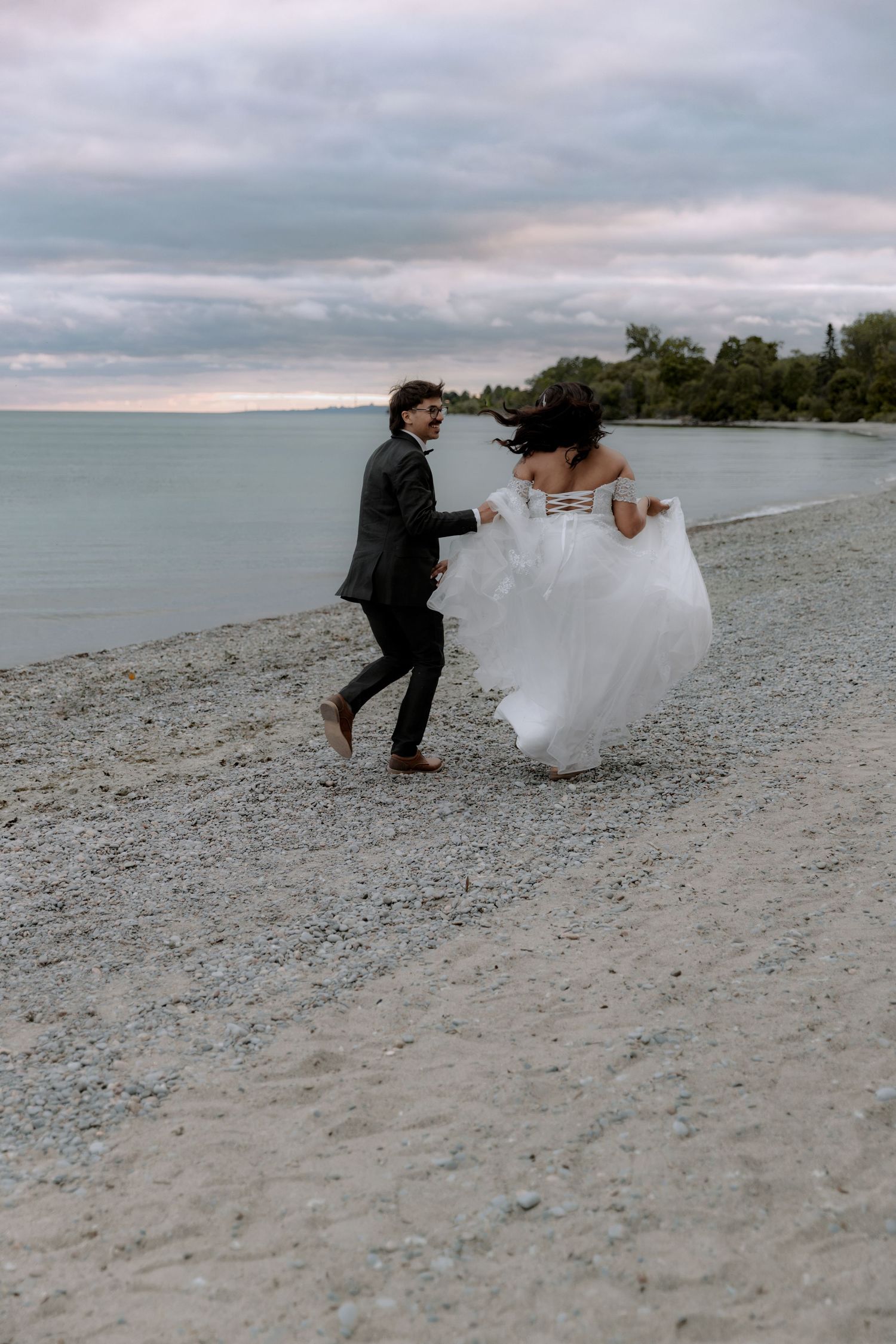 Bride and groom running barefoot on a rocky beach in wedding attire, with ocean and cloudy sky backdrop.