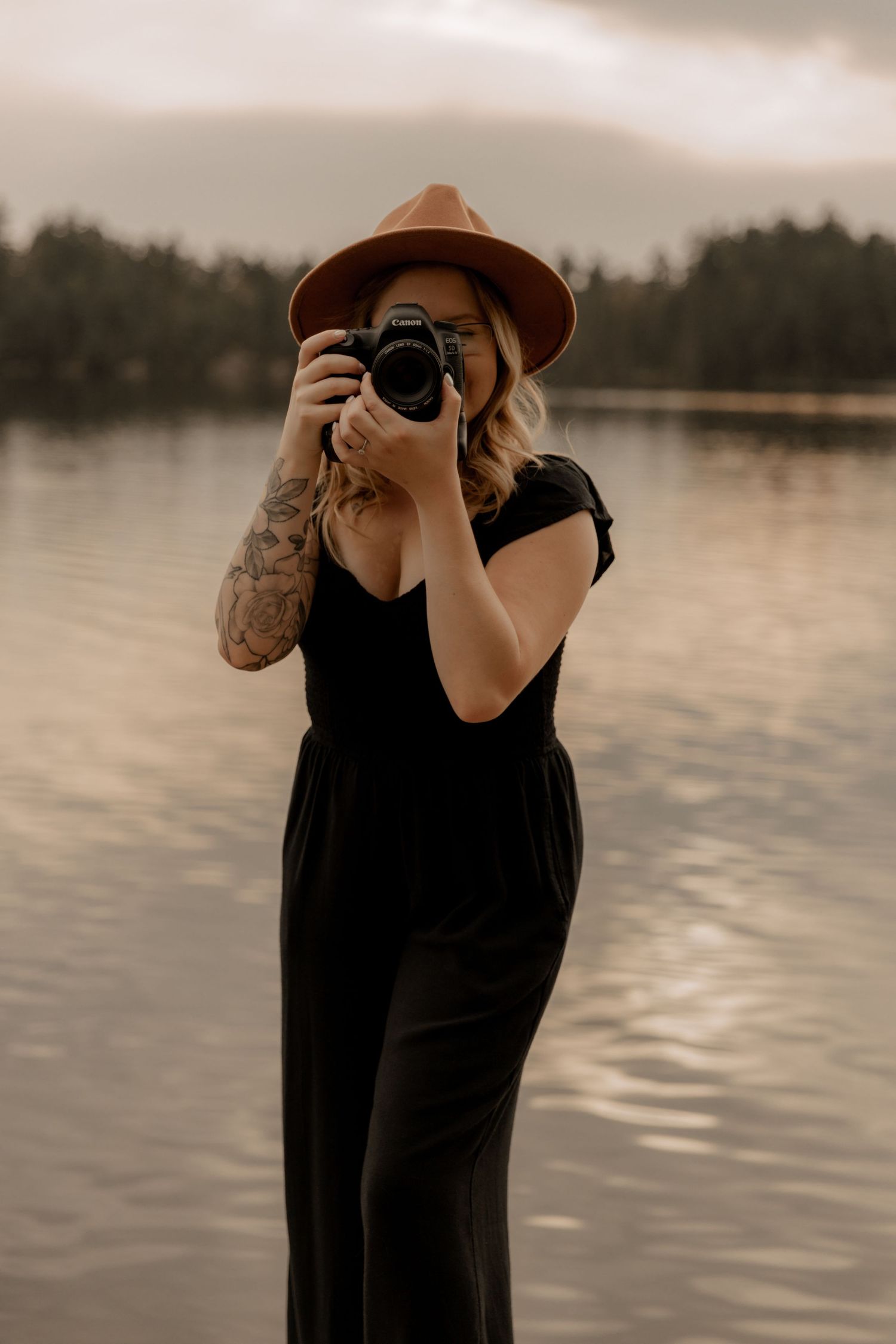 Photographer wearing brown hat and black dress takes pictures by a serene lake on a misty day.