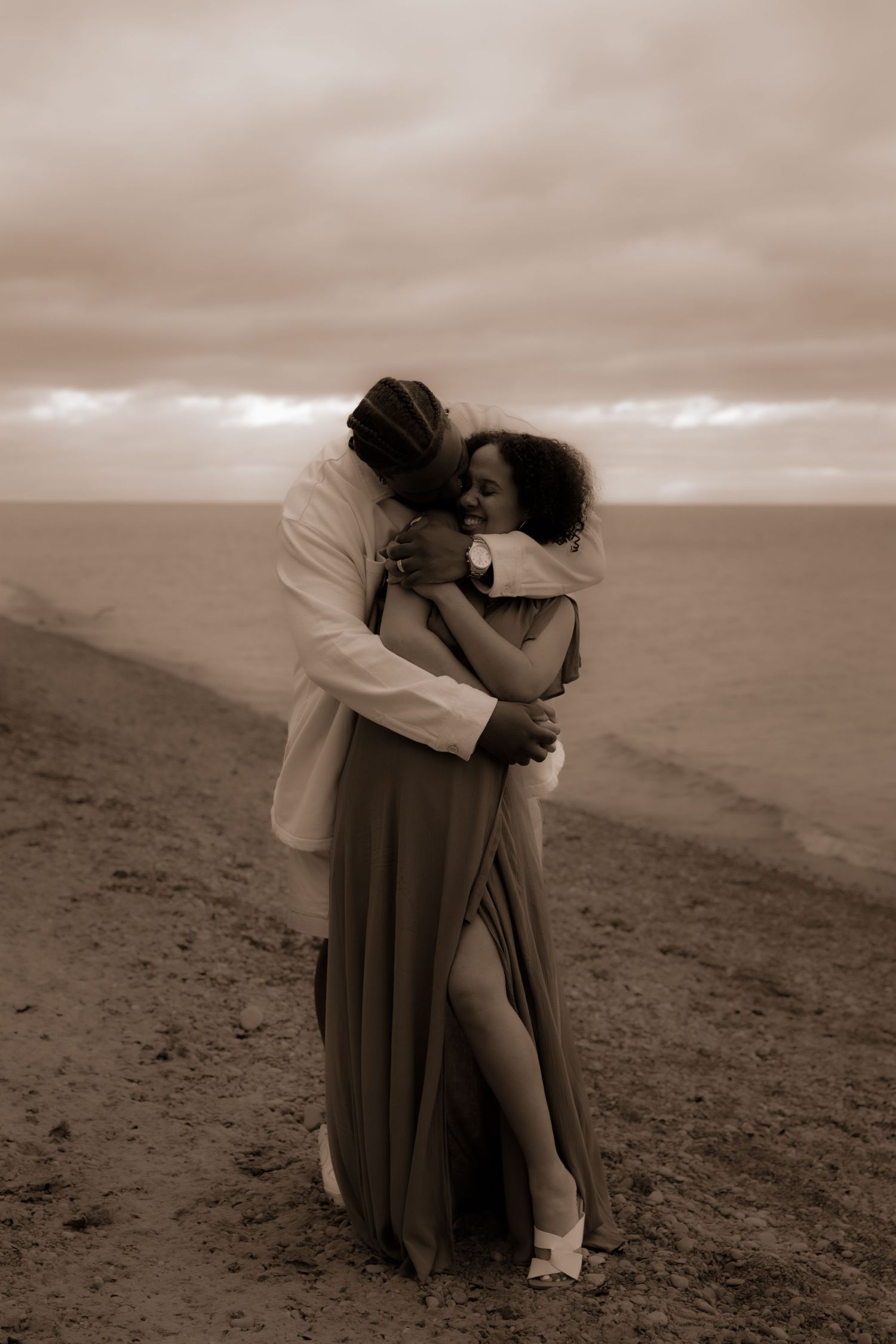 Couple embracing romantically on a beach at dusk, woman in flowing dress, man in white jacket, sepia tone.