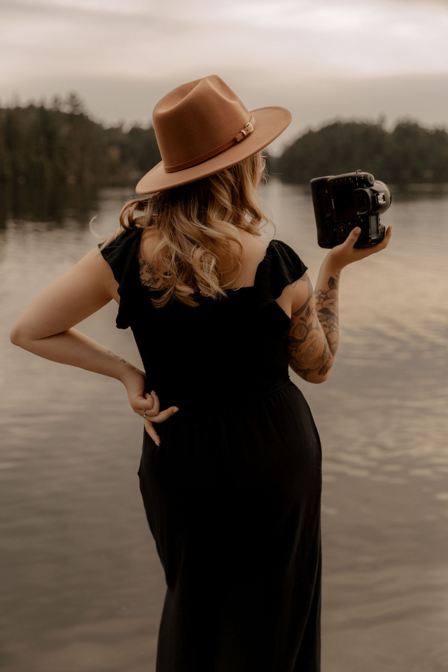 Photographer in black dress and brown hat holds camera while standing by a lake.