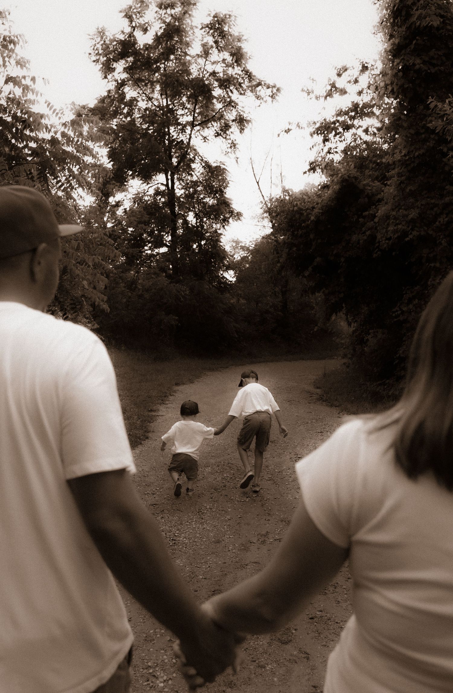 Sepia-toned family walking on a wooded trail, parents holding hands while two children run ahead on a dirt path.