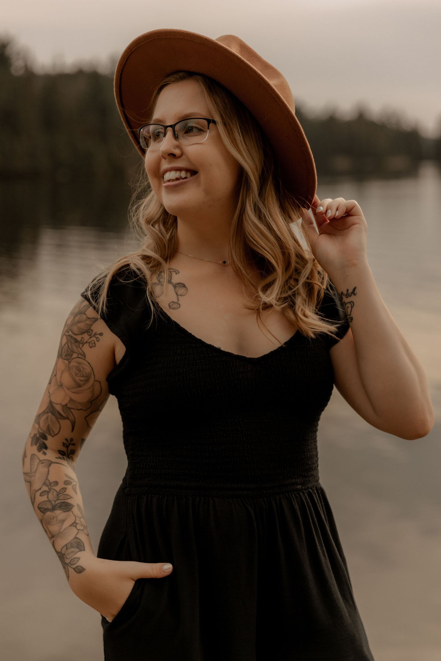 Person in black dress and brown hat smiles while adjusting their hat by a lake.
