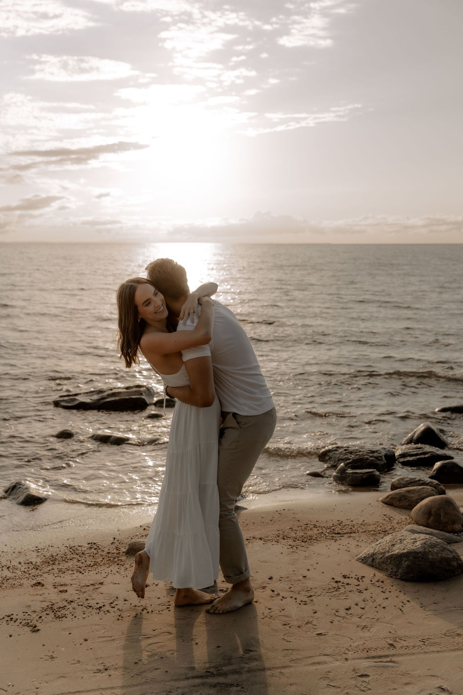 A romantic couple embrace on a rocky beach during sunset.