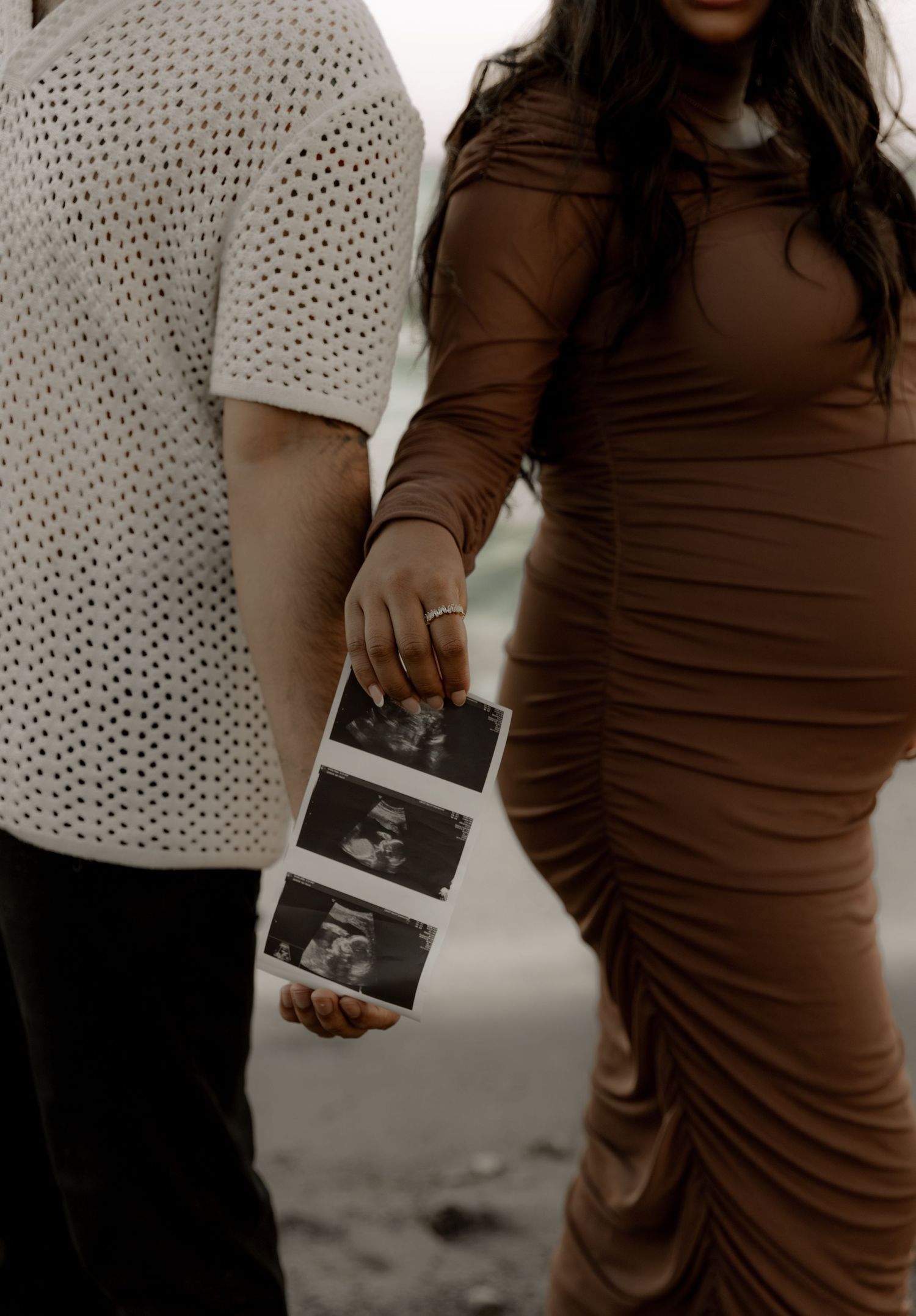 Couple in casual attire holding ultrasound photos on beach during maternity photoshoot.