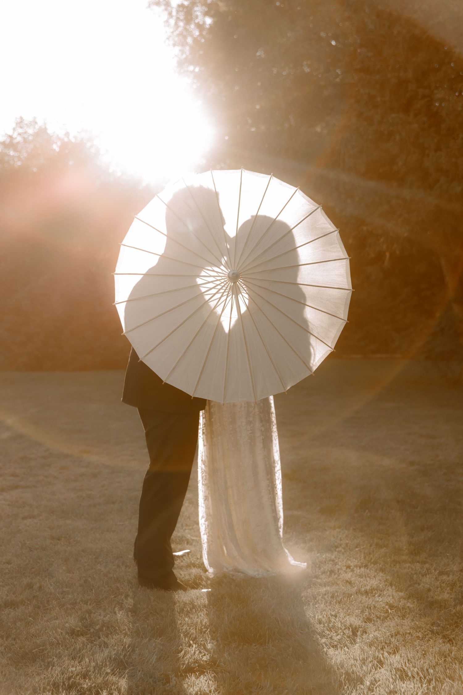 A romantic silhouette of a couple behind a white parasol stands against bright sunlight creating a dreamy sepia-toned scene.