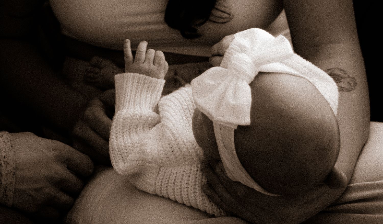 Sepia-toned photo of a newborn baby in white knit outfit and bow headband being cradled while breastfeeding.