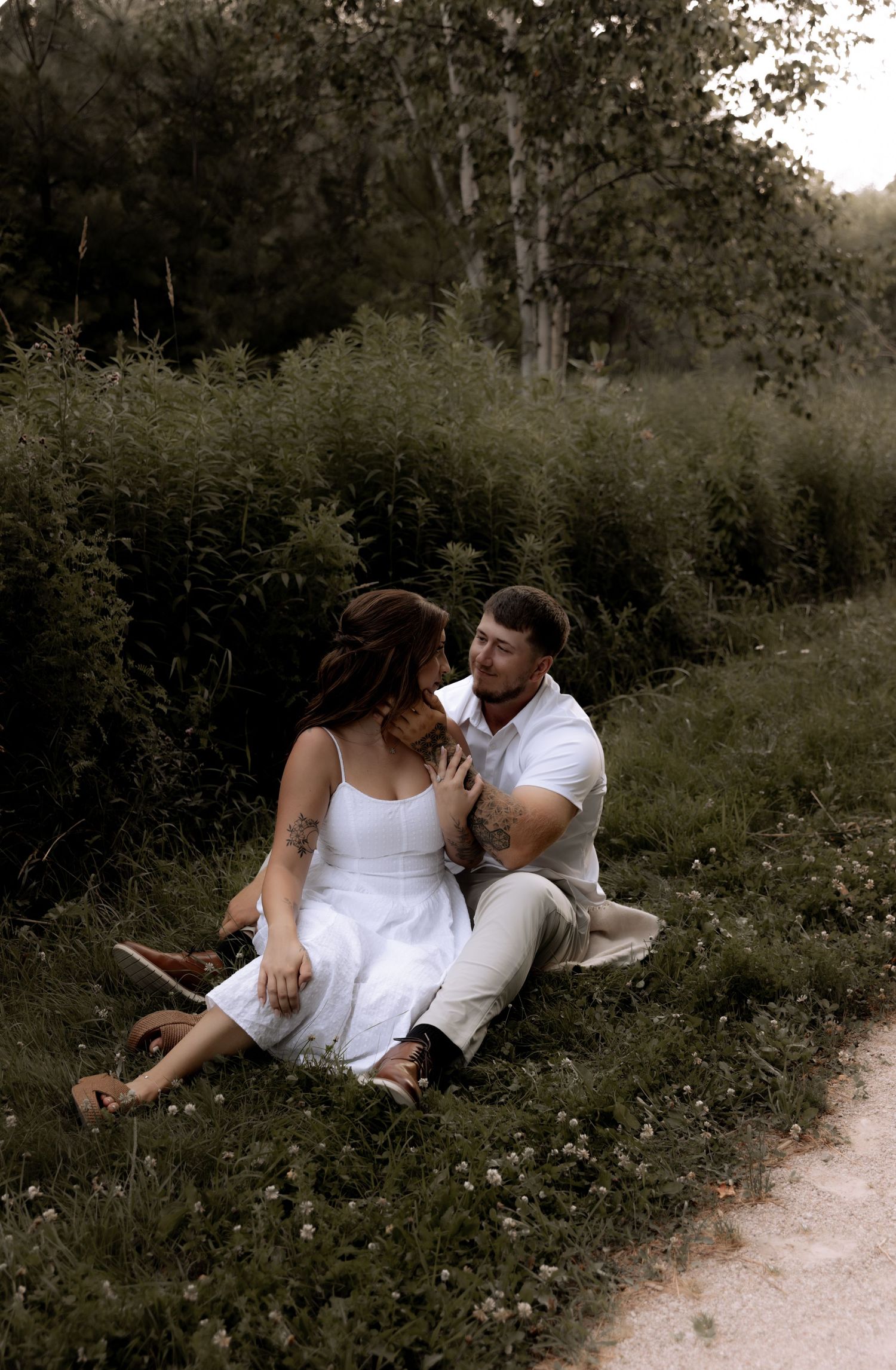 A couple sits together on a grassy hillside, the woman in a white dress leaning toward a man in a white shirt.
