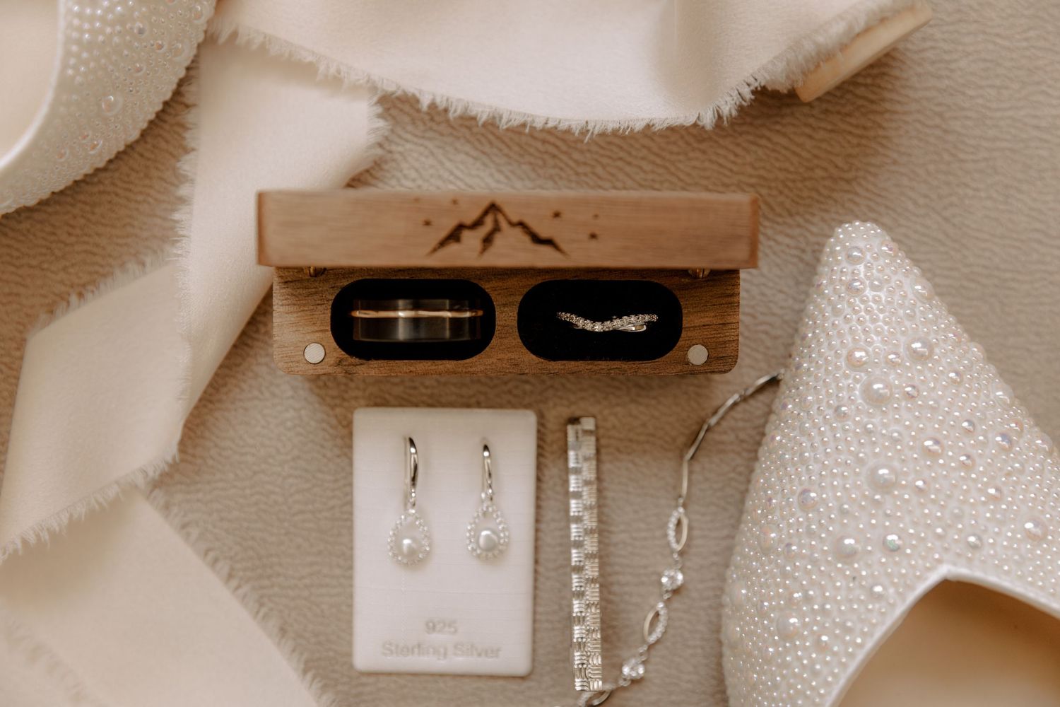 Wedding rings and jewelry laid out with a wooden box featuring mountain engravings on a neutral textured background.