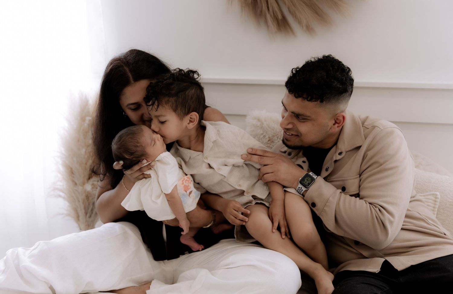 A loving family of four sits together in a bright white room, with parents cuddling two young toddlers.